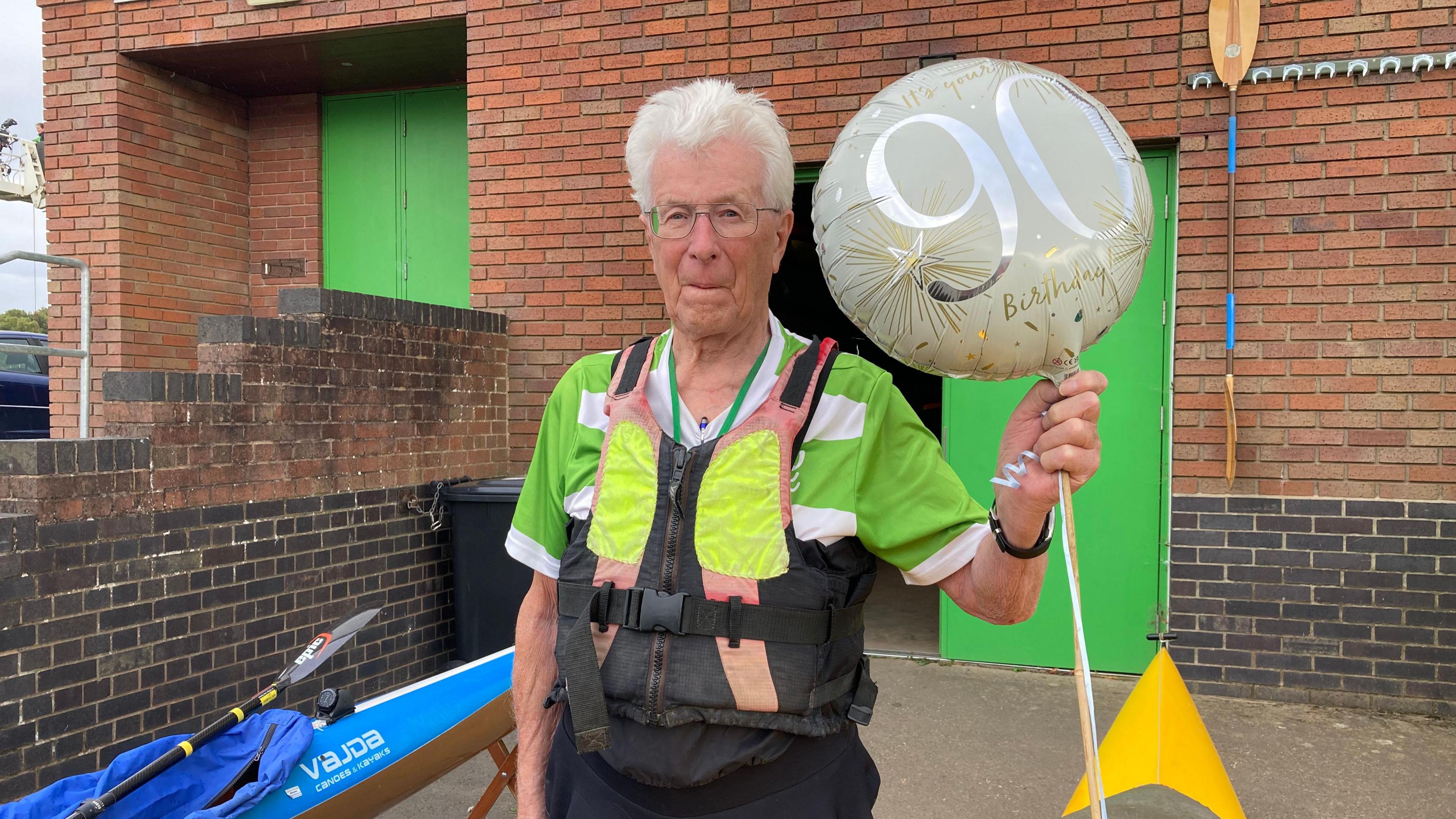 Man wearing a black, orange and bright yellow life jacket holds a gold and silver 90th birthday balloon with a blue canoe and yellow paddle in the background 