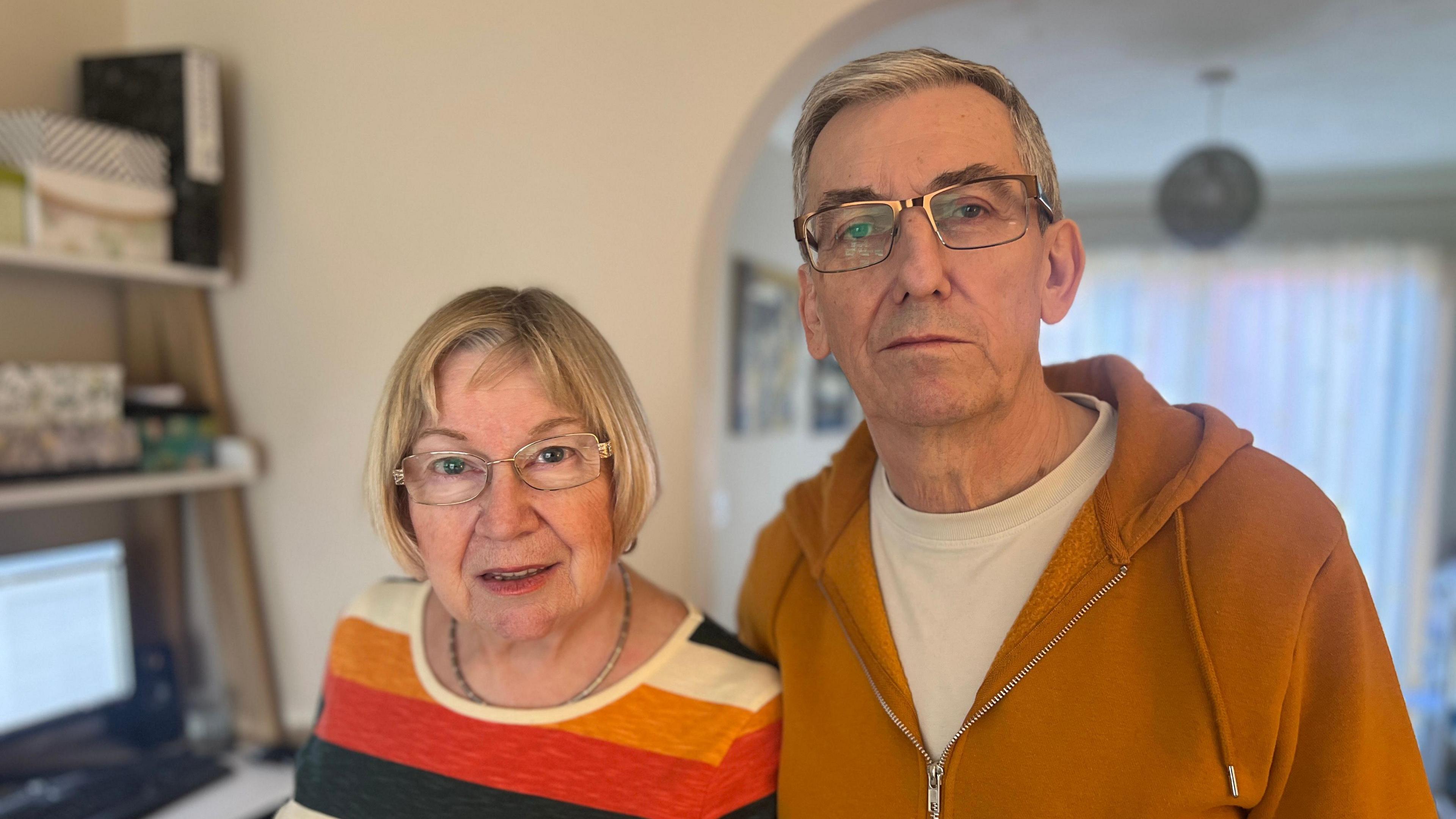 Alyson and Chris Austin stand in the living room of their home. They are both wearing orange-toned jumpers, and are looking at the camera. Alyson has short blonde hair and Chris has cropped silver hair, and they are both wearing glasses.