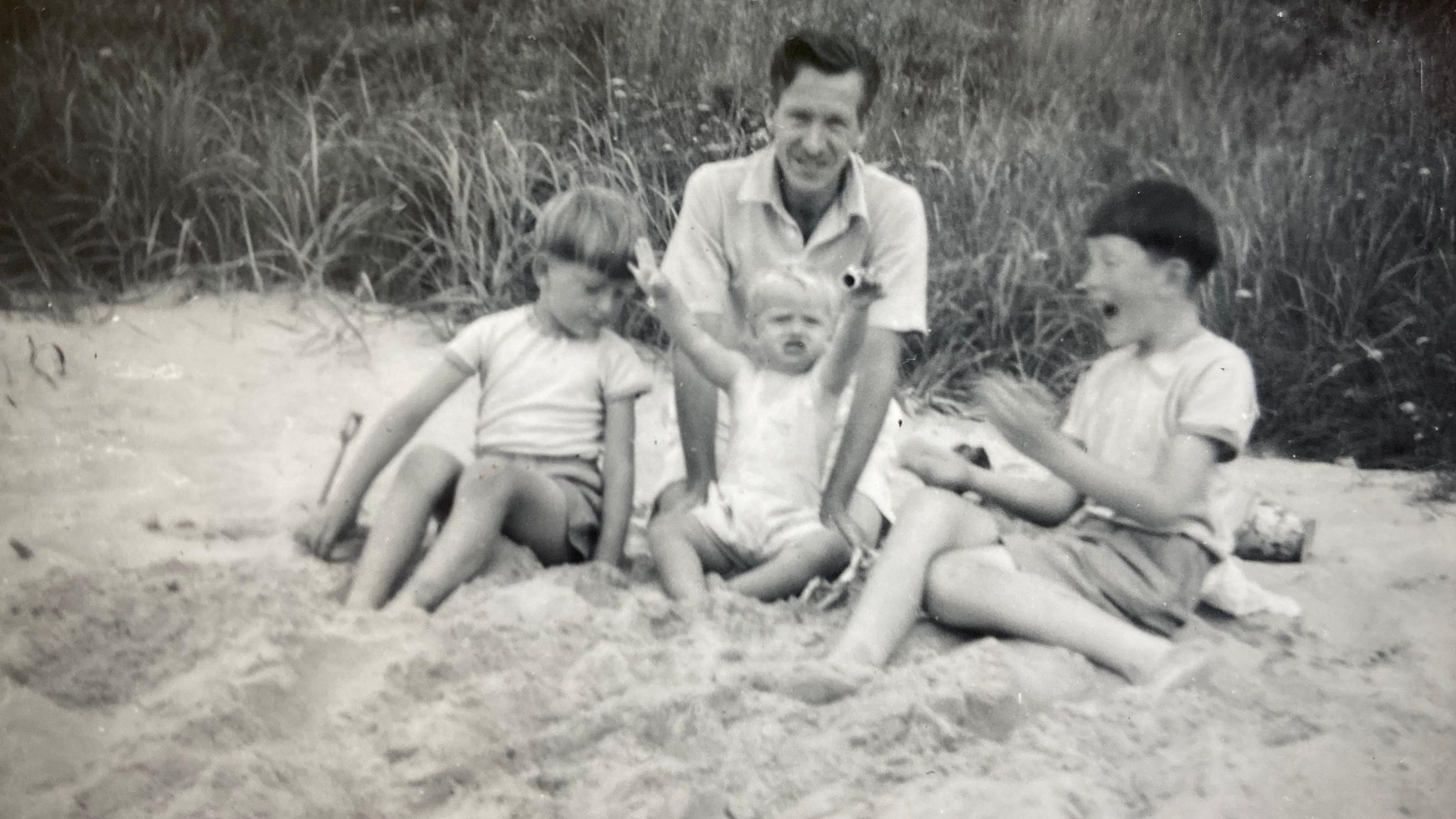 Leslie on a beach with his three children, Richard, Mary and Michael. Leslie is looking at the camera, and Mary has her arms up, with Michael looking at her and clapping. The image is black and white. 