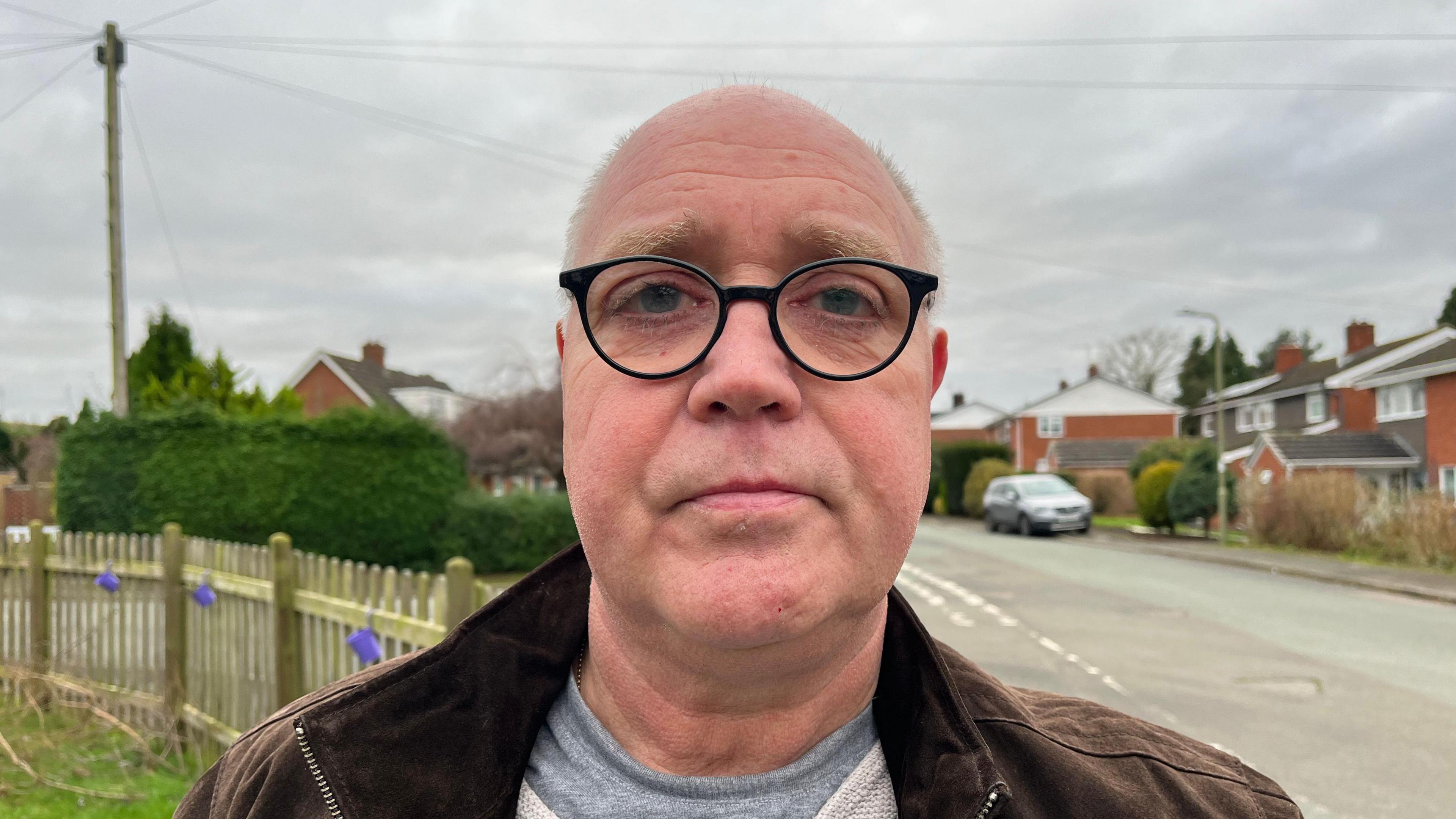 Paul looking down the camera, he's wearing a brown jacket with a grey top underneath. Behind him you can see Pinewood Road, a few houses and trees are visible in the background. The sky is grey and overcast.