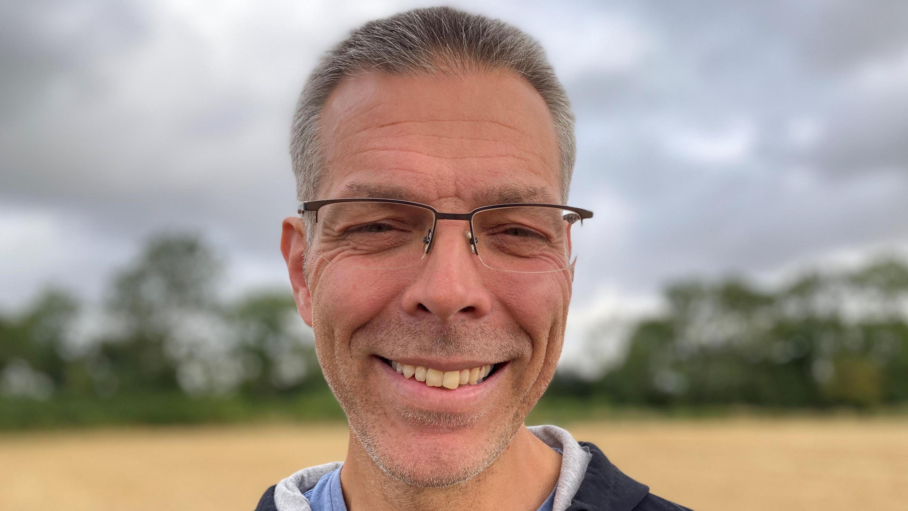 A head and shoulders photograph of Ian Keel. Standing in a field, he is smiling for the camera and has short grey hair, glasses and is wearing a blue hoody.