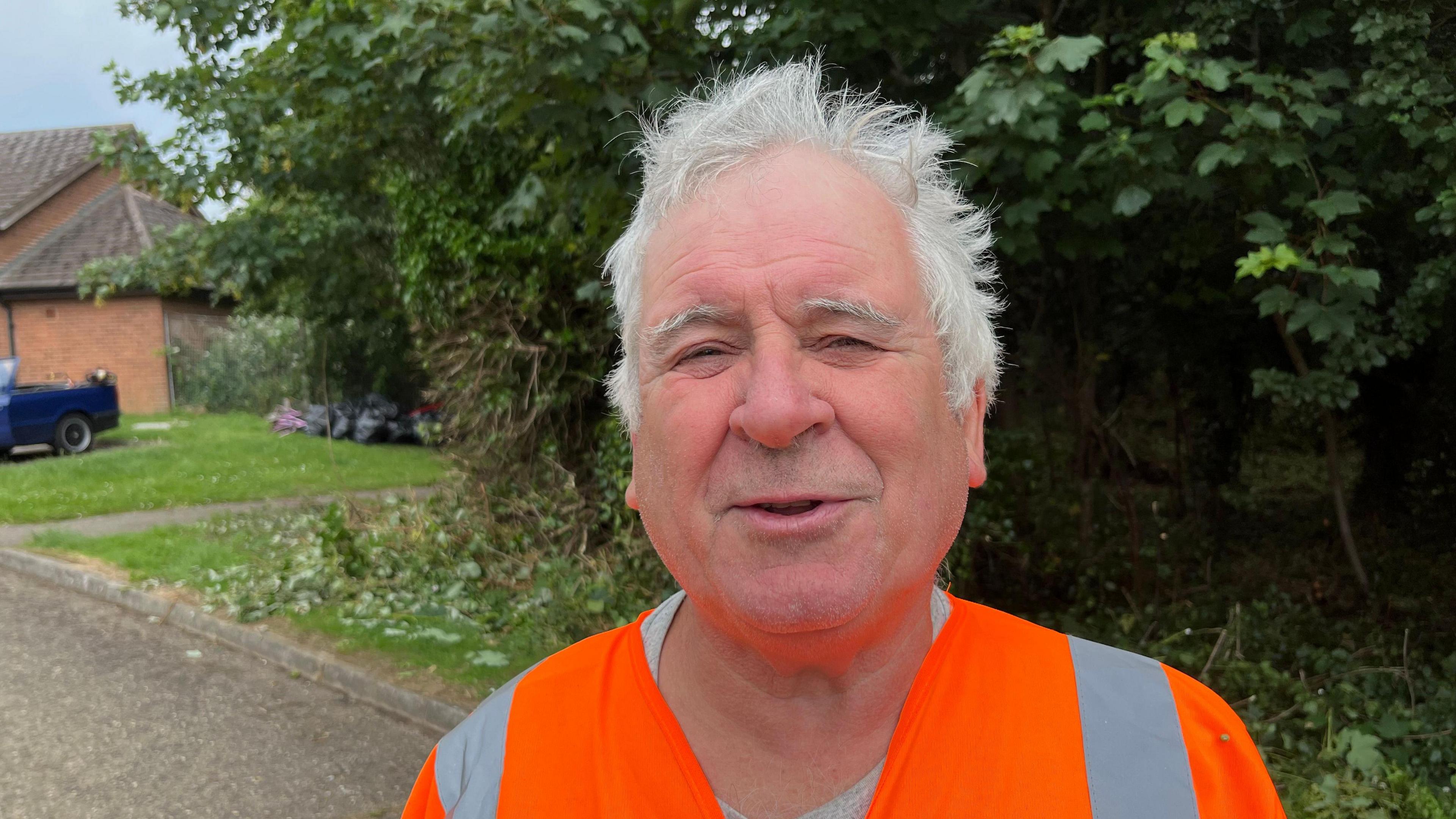 A man is looking at the camera. He has grey hair and is wearing a hi-vis orange jacket with a grey jumper underneath. There are trees and bushes behind him.
