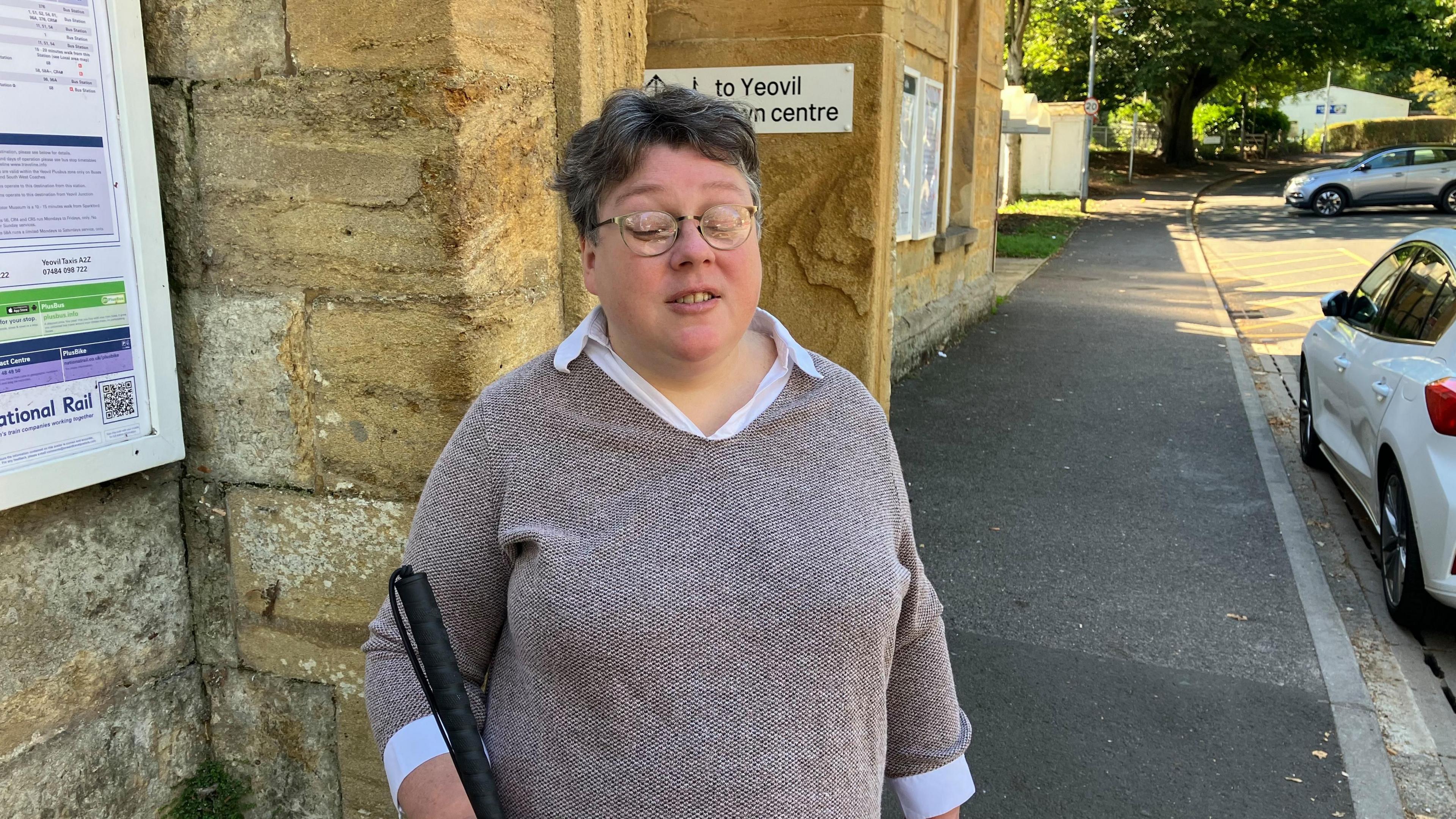 A woman wearing glasses holding a white cane standing outside a yellow stone train station building