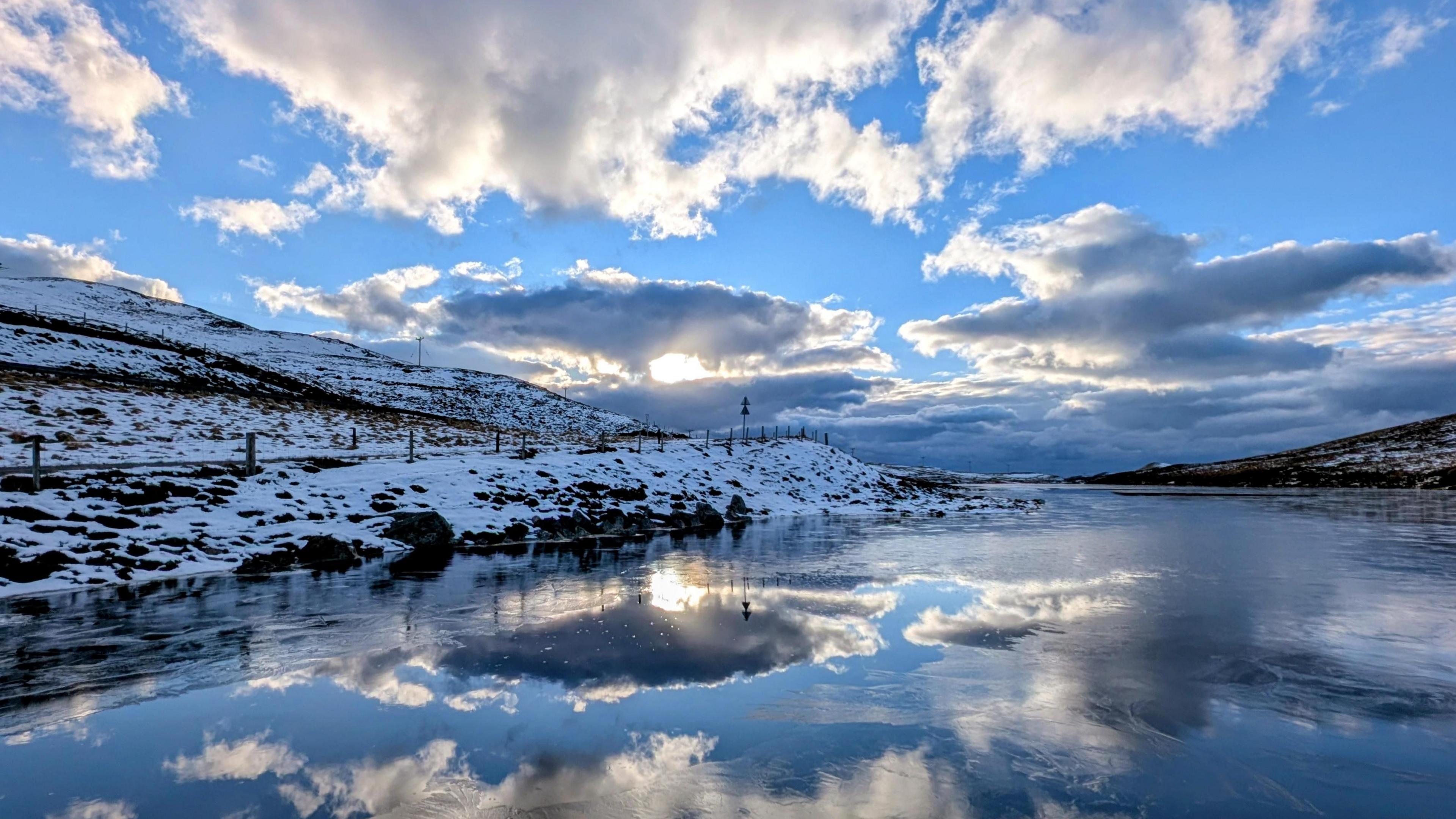 A photo of a snowy hillside beside a frozen lake with a partly cloudy sky overhead and the sunrise in the background