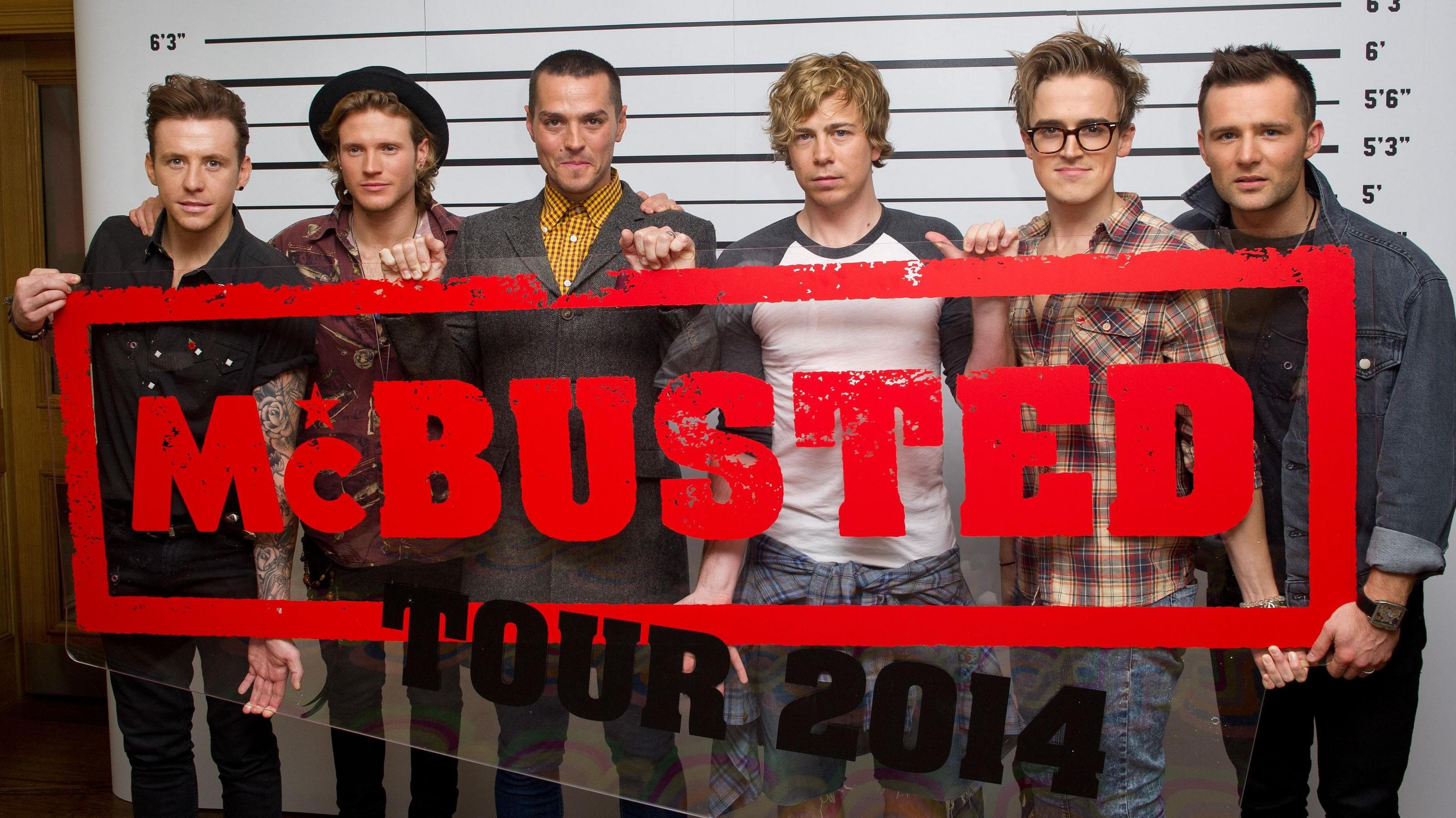 McBusted holding a sign that says "McBusted Tour 2014". On the left is Danny Jones, on his right is Dougie Poynter, then Matt Willis, then James Bourne, then Tom Fletcher, then Harry Judd. They are all stood in front of a height chart.