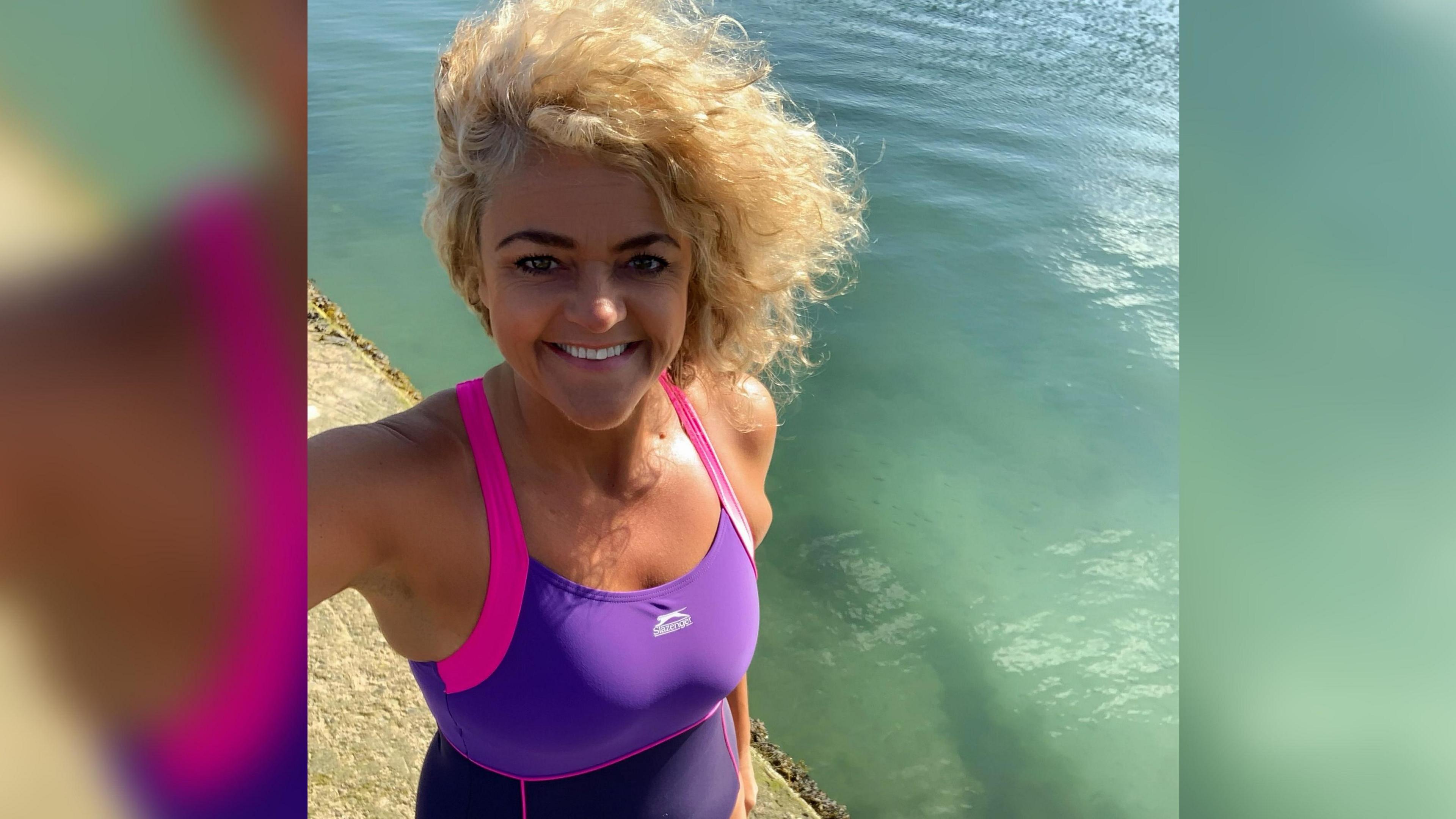 Rebekah Kefford in a selfie standing next to the sea with the sun shining off it. She has blond curly hair, is wearing a pink and purple swim suit and is smiling at the camera.