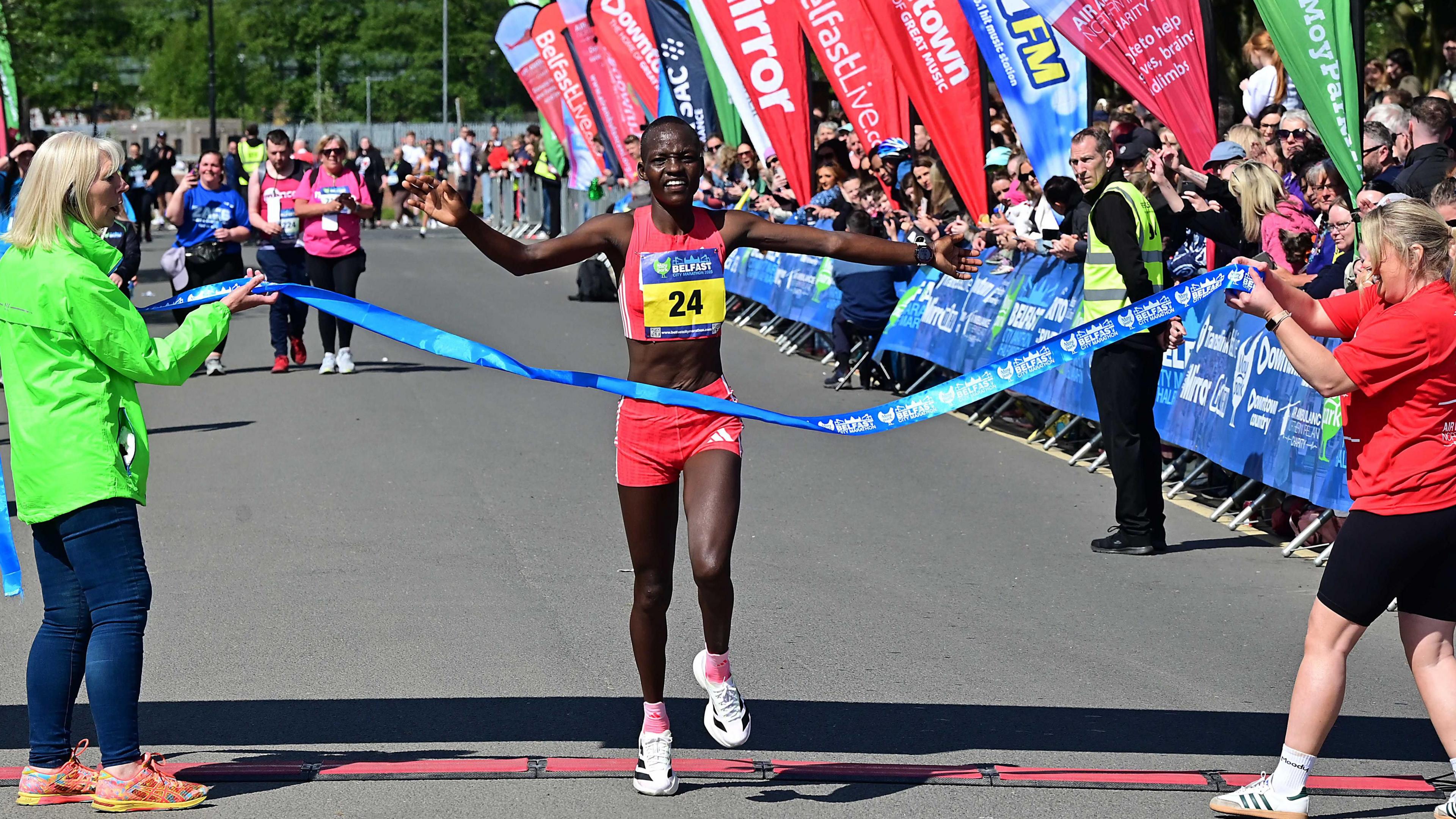 A lady with short black hair crossing the finish line in the marathon. She is wearing a pink top and shorts and white trainers. She is running through a blue ribbon being held up by two women who have blonde hair. Behind her are spectators.