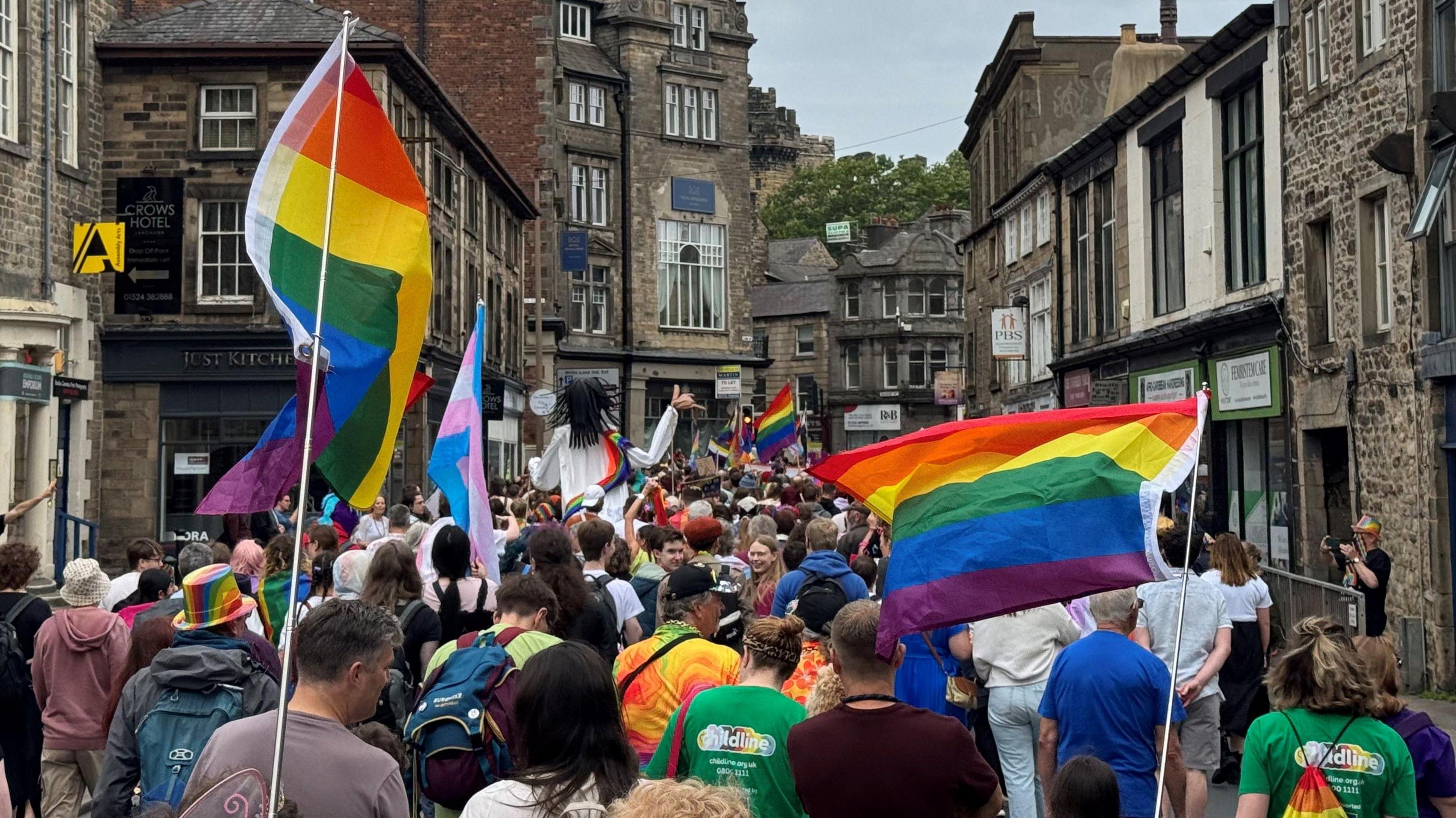 People fly rainbow pride flags as they stream through a historic narrow street in Lancaster city centre with old shops lining both sides of the road