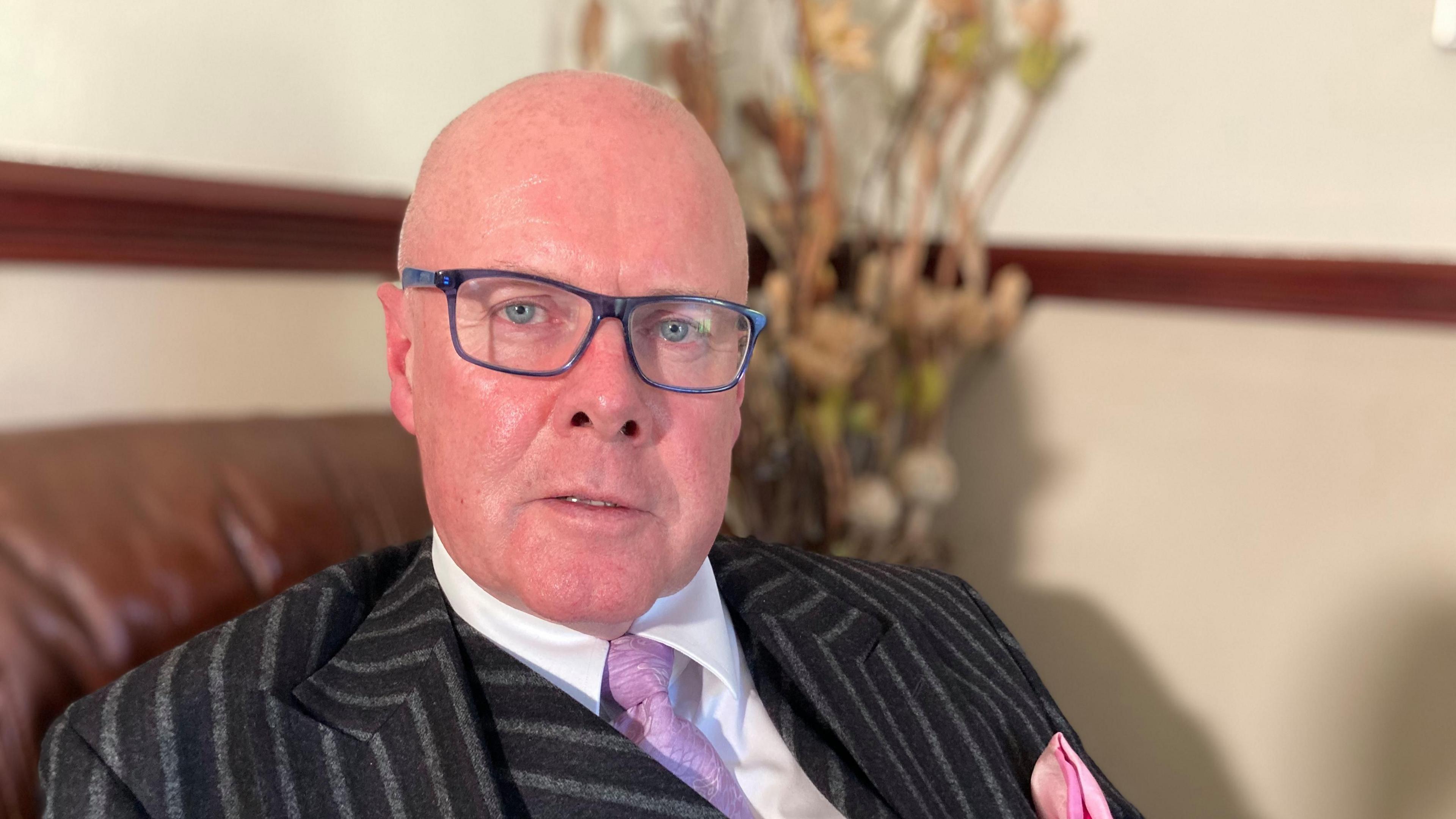 Michael Rushe is looking at the camera and is wearing a pair of square blue framed glasses. He is wearing a black and grey striped blazer and waistcoat over a white shirt and pink tie, with a matching pink pocket square. Michael is sat on a brown leather armchair. In the background can be seen a magnolia wall with a dark brown dado rail and just behind his head is some dried foliage. 