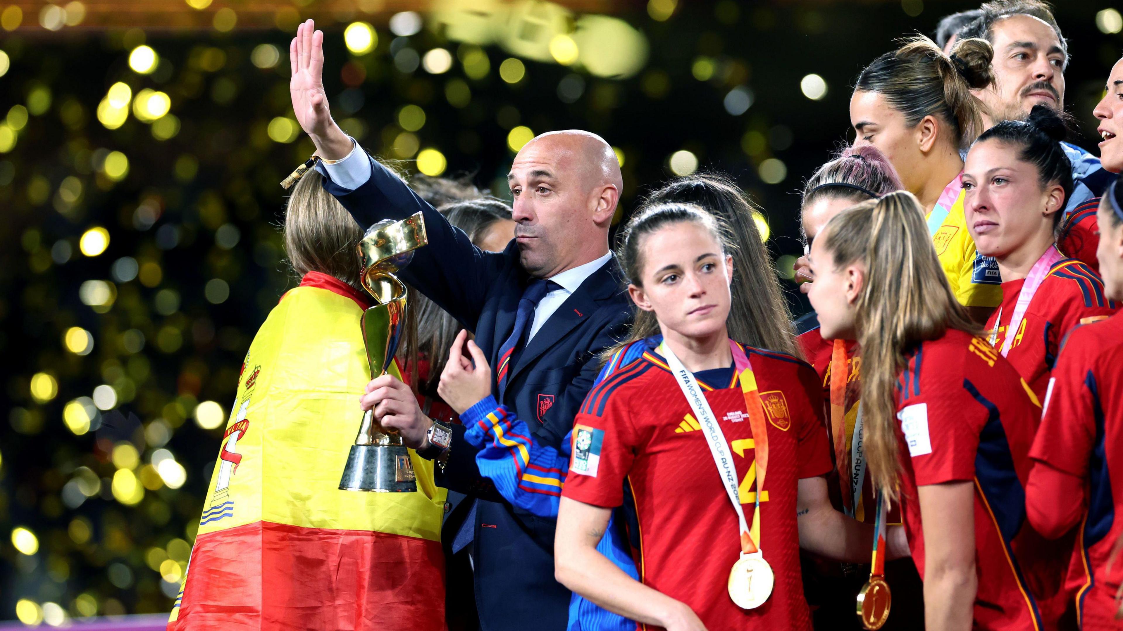 Rubiales holding the Women's  Football World Cup Trophy on the day of the final. He is surrounded by the players who are all looking away from him while he waves to the crowd. 