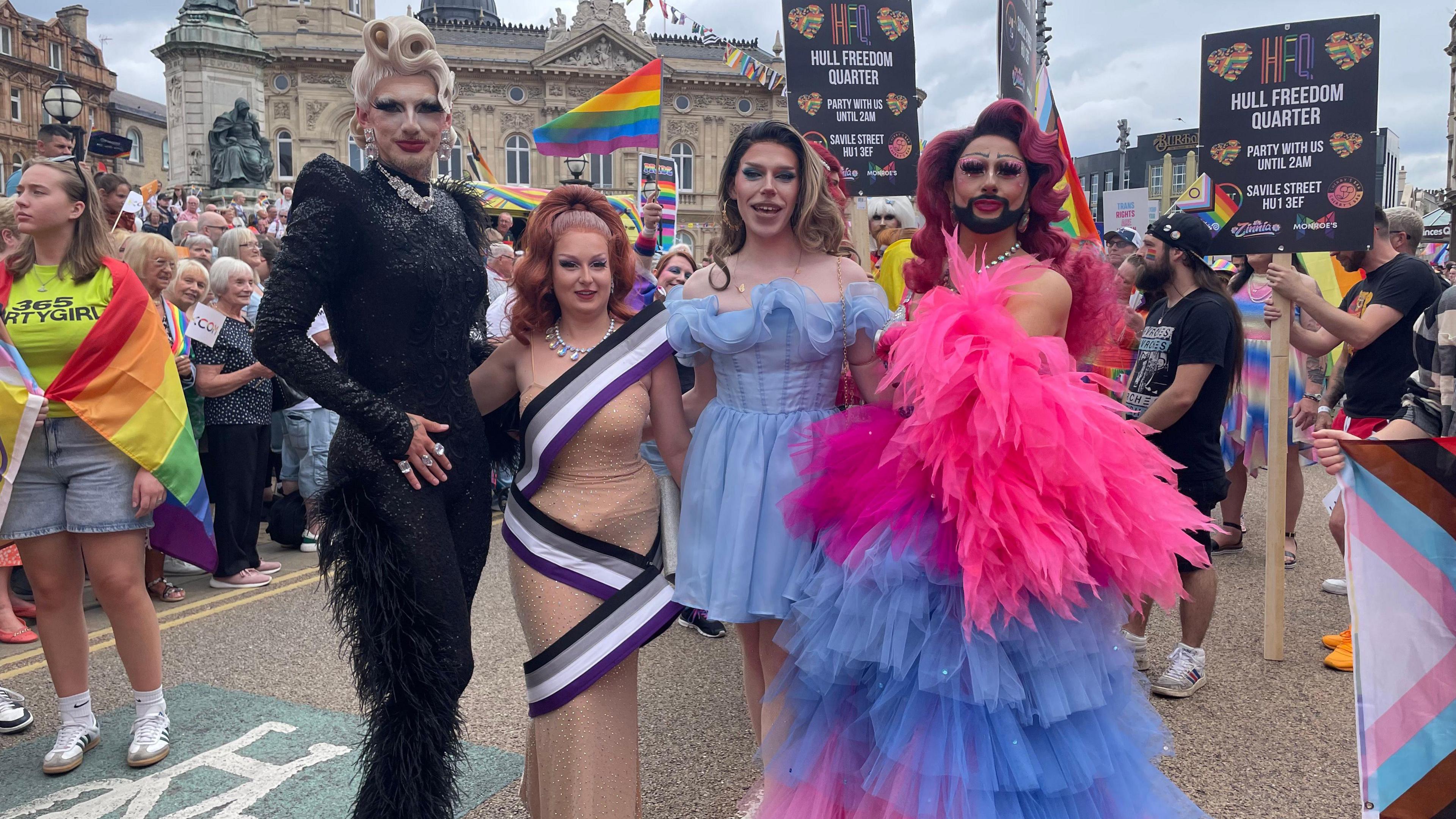 Four drag queens posing for a photo. One has a beard and a netted frock with multiple frills. All four have wigs and plenty of make-up on.