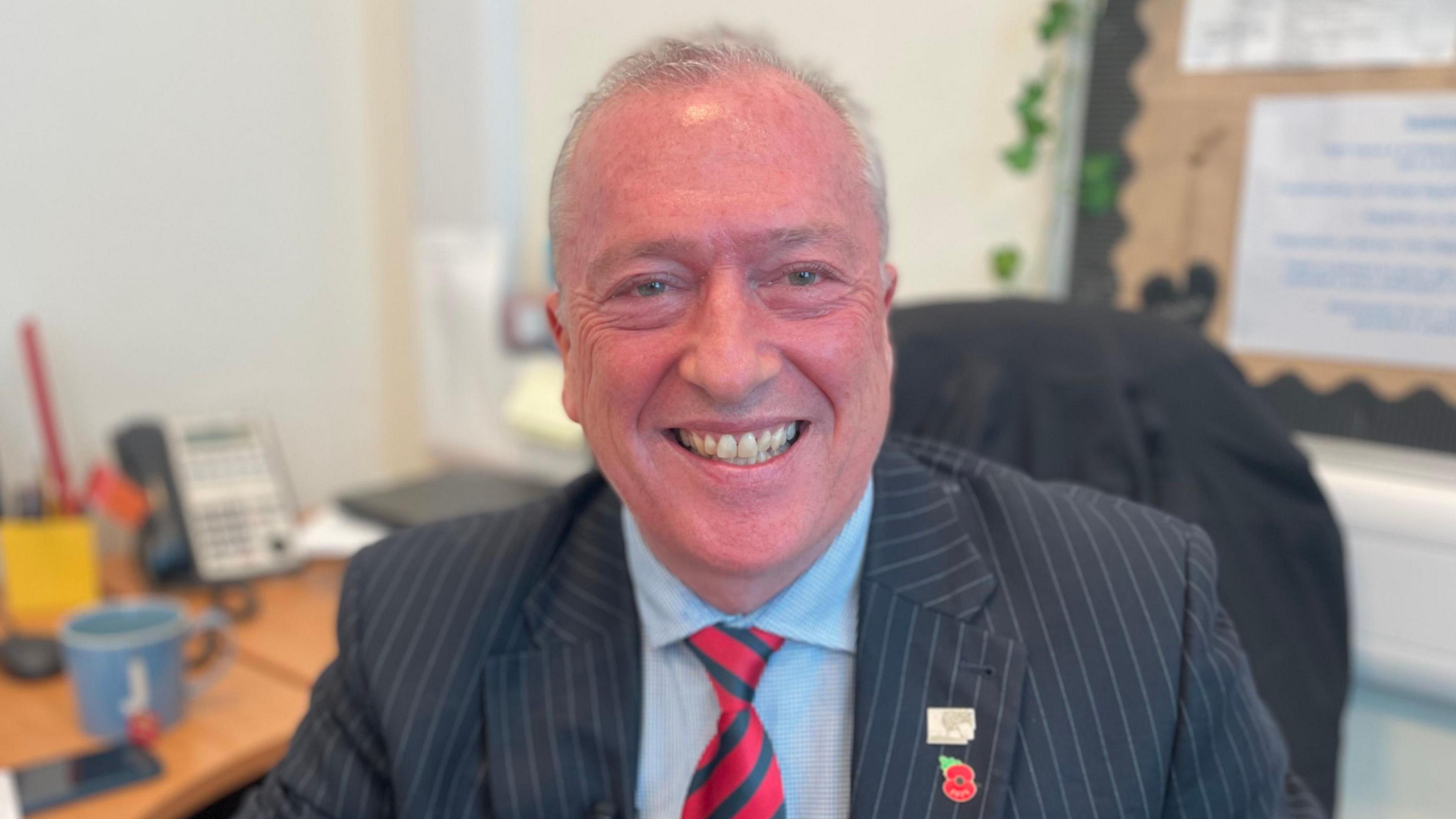 Dyn gyda gwallt llwyd byr yn gwenu ac yn gwisgo siaced lwyd pinstripe a thei streips coch a gwyrdd tywyll. Mae'n eistedd mewn swyddfa gyda desg tu ol iddo.