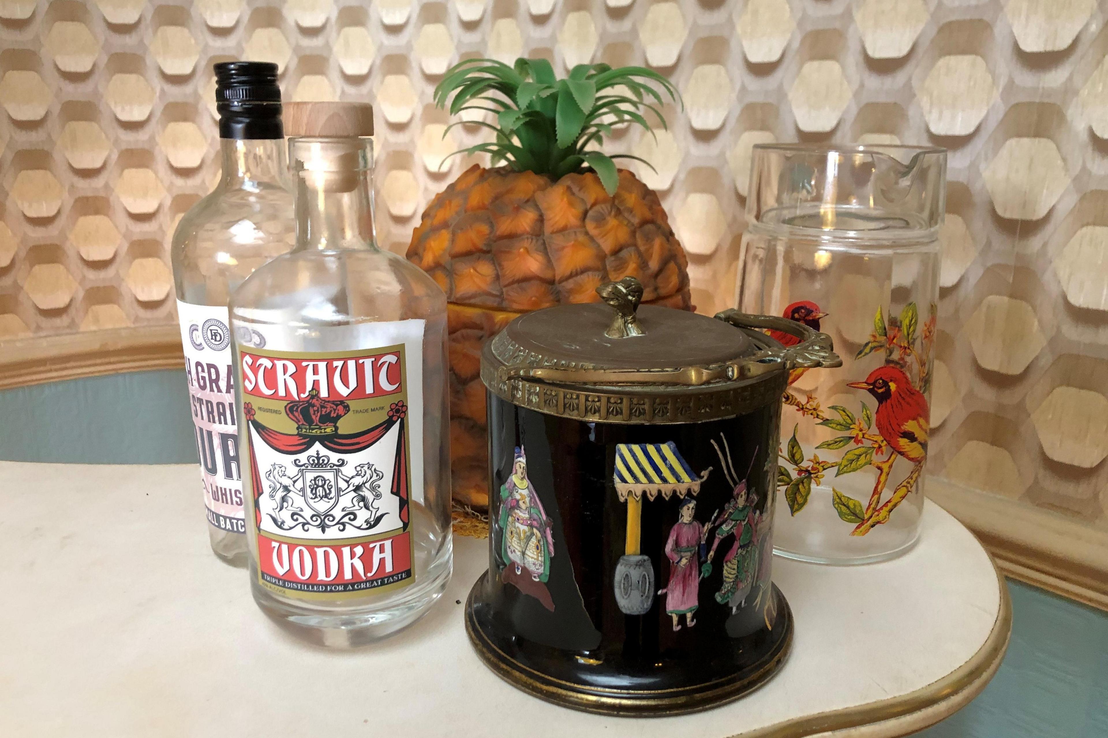 Two bottles of alcohol, a jug with birds on it, a plastic pineapple and a black metal ice box with people painted on it sitting together on a surface counter