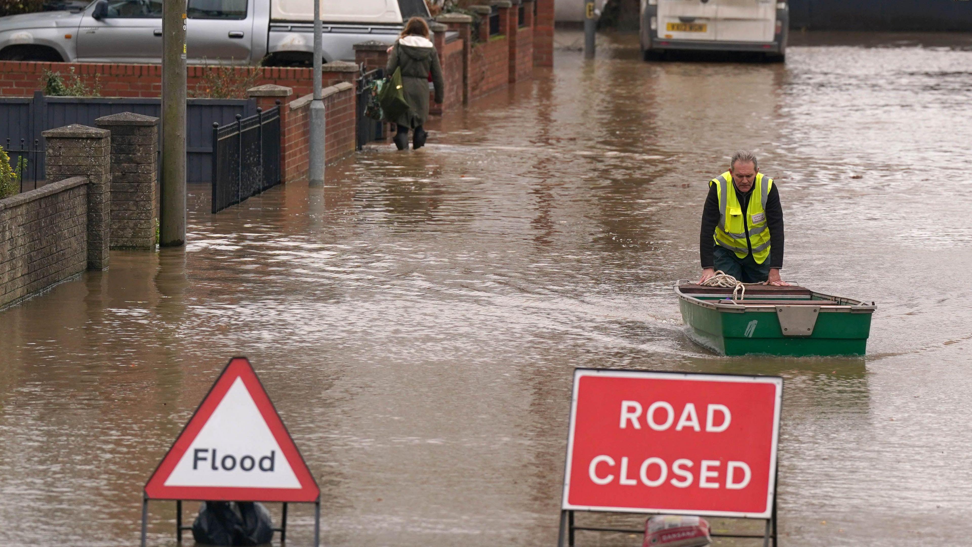 A man in a high-vis jacket pushes a small boat through murky-looking water. In front of him there are tow signs, both are red and white. The sign on the left says 'Flood', while the sign on the right says 'ROAD CLOSED'. In the background, another person is walking through the water in the opposite direction. The water is roughly knee height, and is on what appears to be a residential street. 