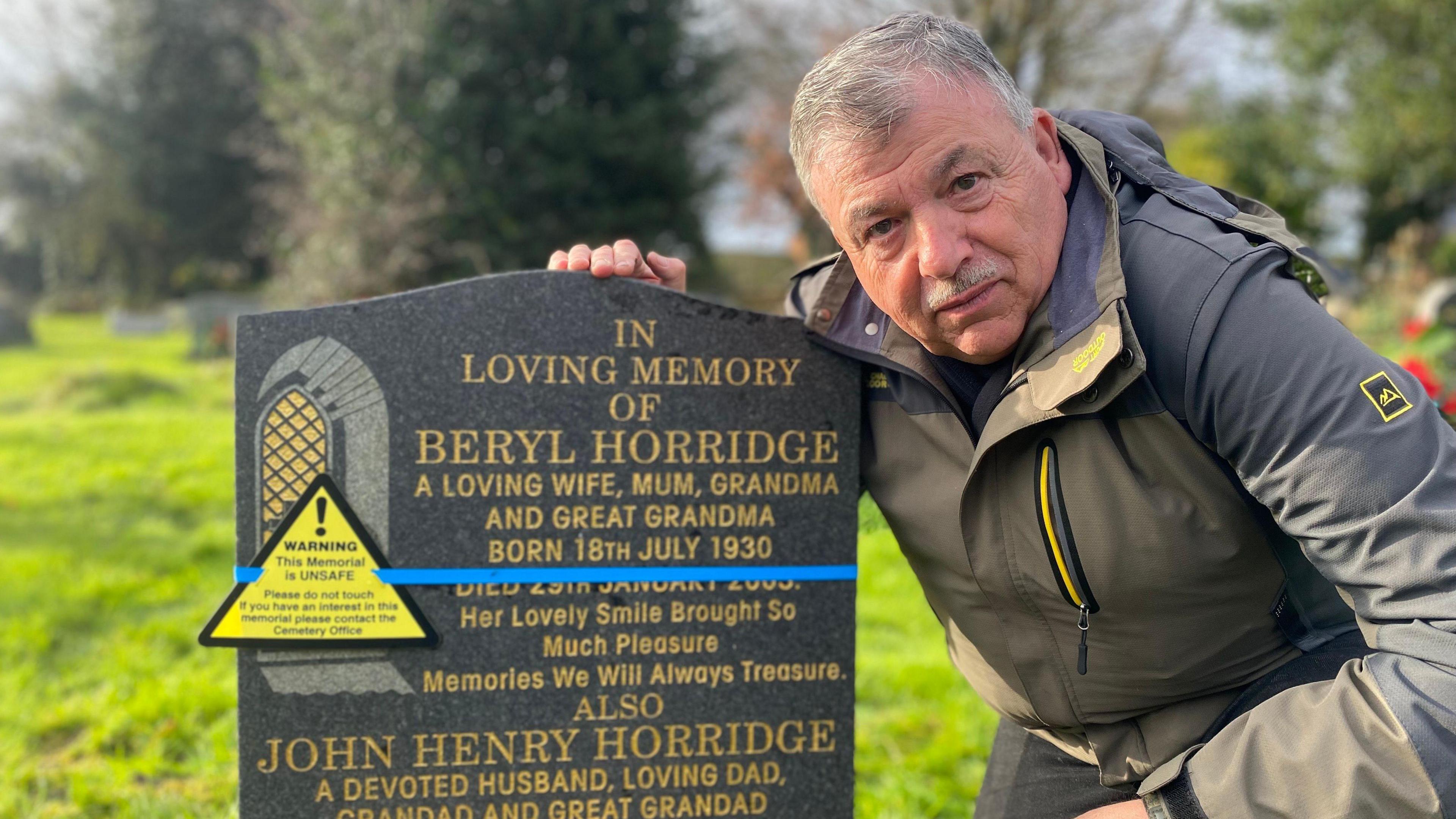 A man crouching down next to a gravestone with gold writing. The stone has a yellow warning sticker on to mark it as unsafe. The man is wearing a grey and khaki coat.