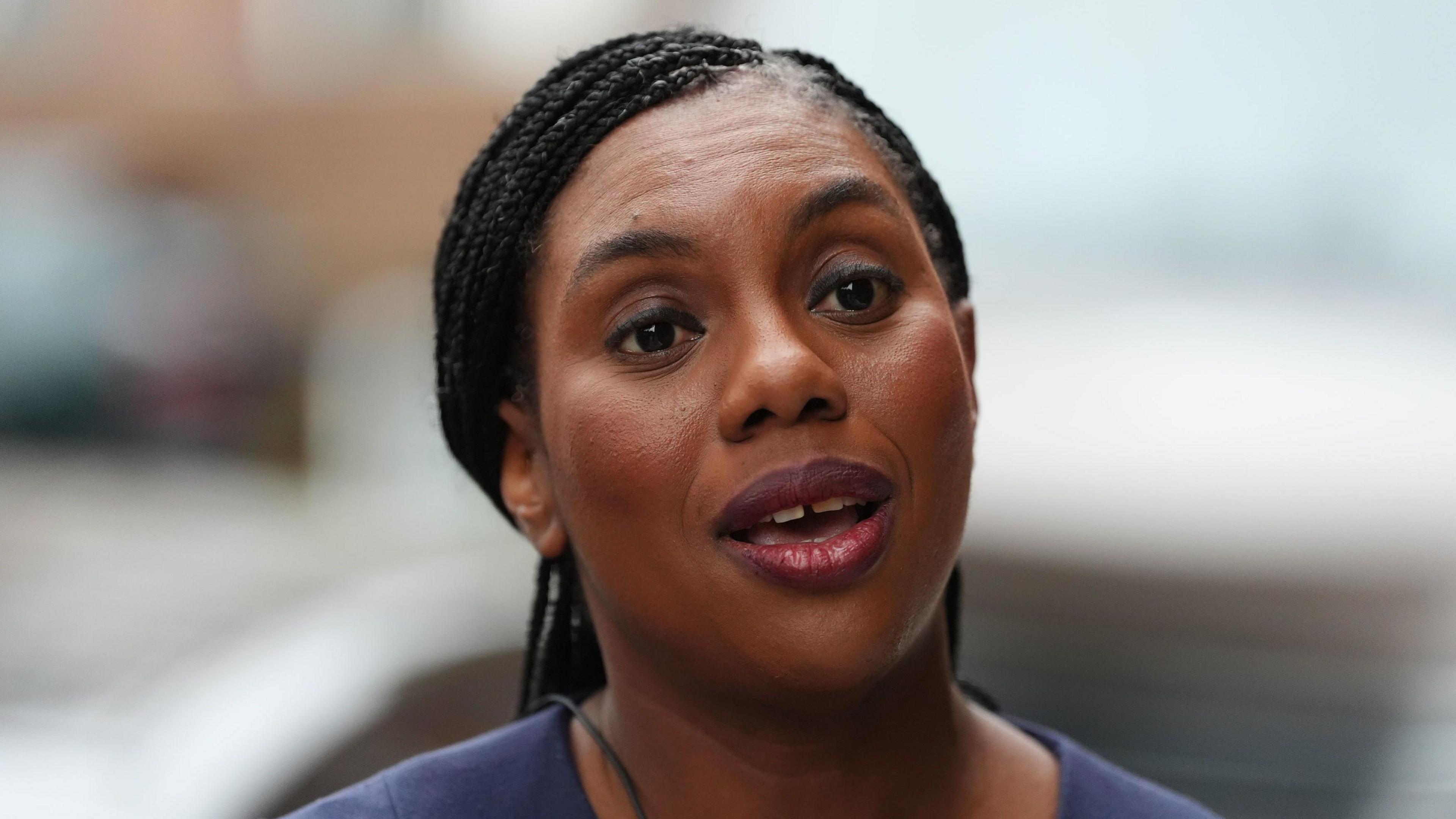 A head and shoulders shot of Kemi Badenoch speaking to someone off camera, looking straight ahead of her. She is wearing a dark purple top.