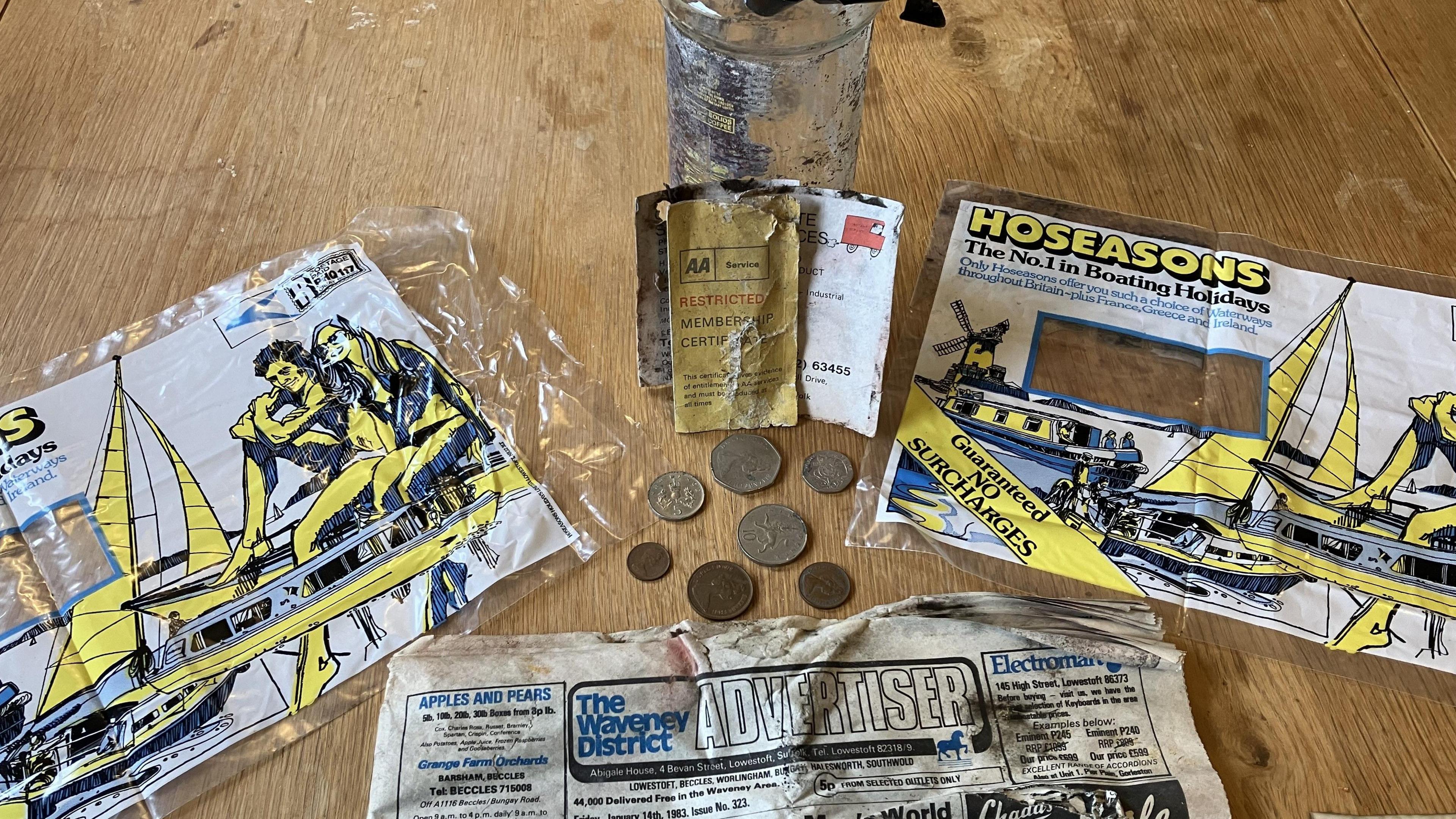 Items from the time capsule are displayed on a wooden table including coins from the era, The Waveney District Advertiser newspaper, an AA member's card and Hoseasons brochure plastic coverings 