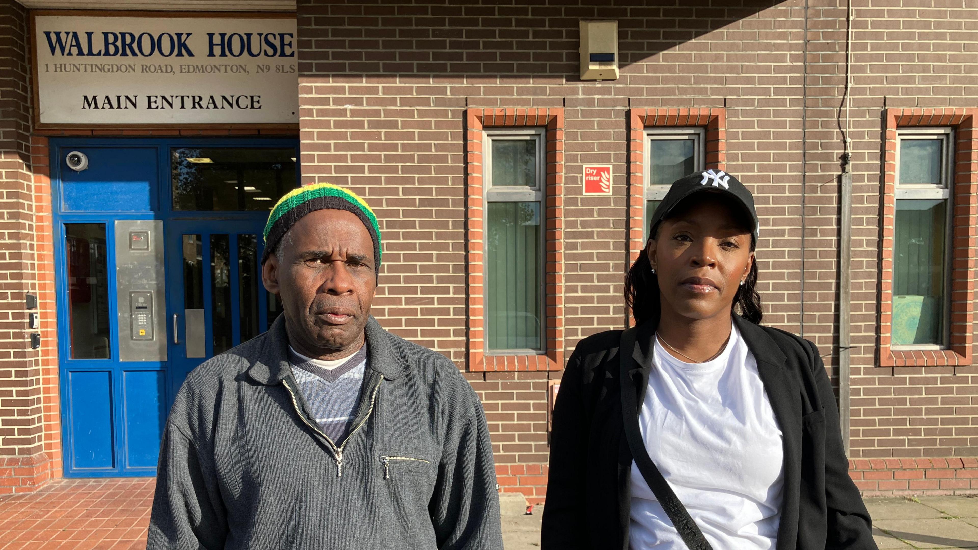 Lesta Hall, a man wearing a red, yellow, blue and black beanie and grey sweatshirt, and Rochelle Wright-Clarke, a woman with long dark hair wearing a black and white baseball cap, white t-shirt and black blazer. They are standing in front of the entrance to Walford House which has blue double doors, a black and white sign and brick walls