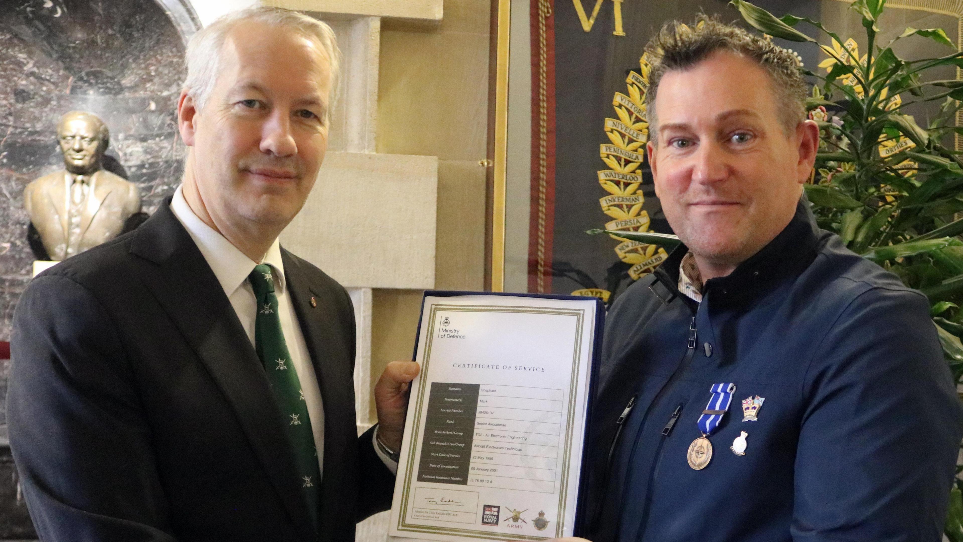 two men looking at camera holding a certificate with a statue and a plant in the background