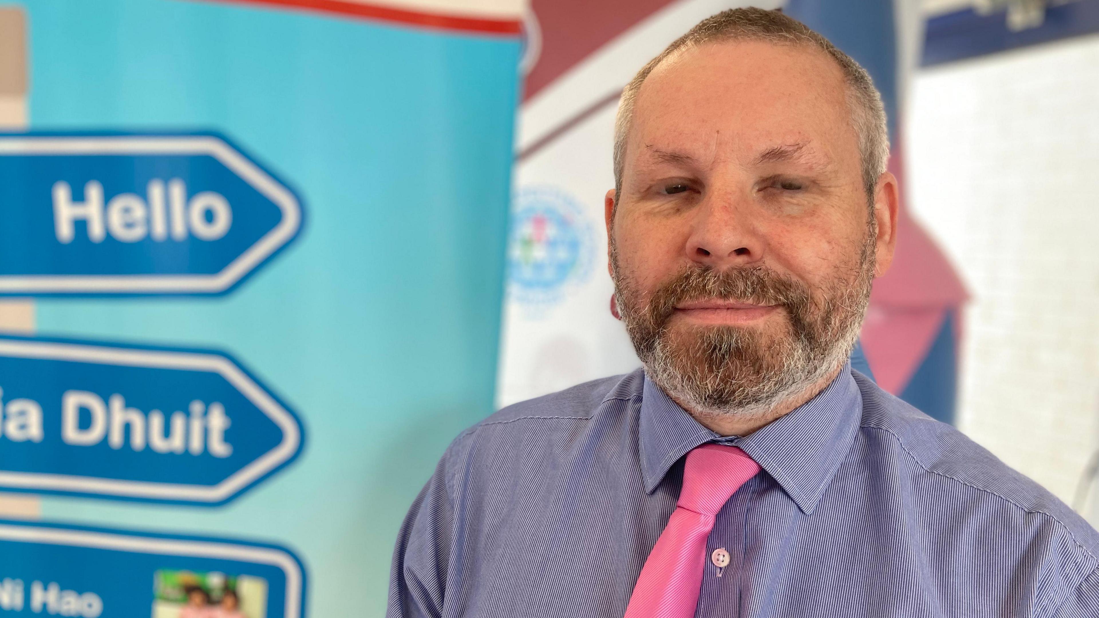 Sean McNamee, who has short grey hair and a beard, stands in front of a colourful poster board with 'hello' and 'dia dhuit' written on blue road signs. Mr McNamee is smiling at the camera and his eyes are slightly closed. He is wearing a blue and white patterned shirt and a pink tie.