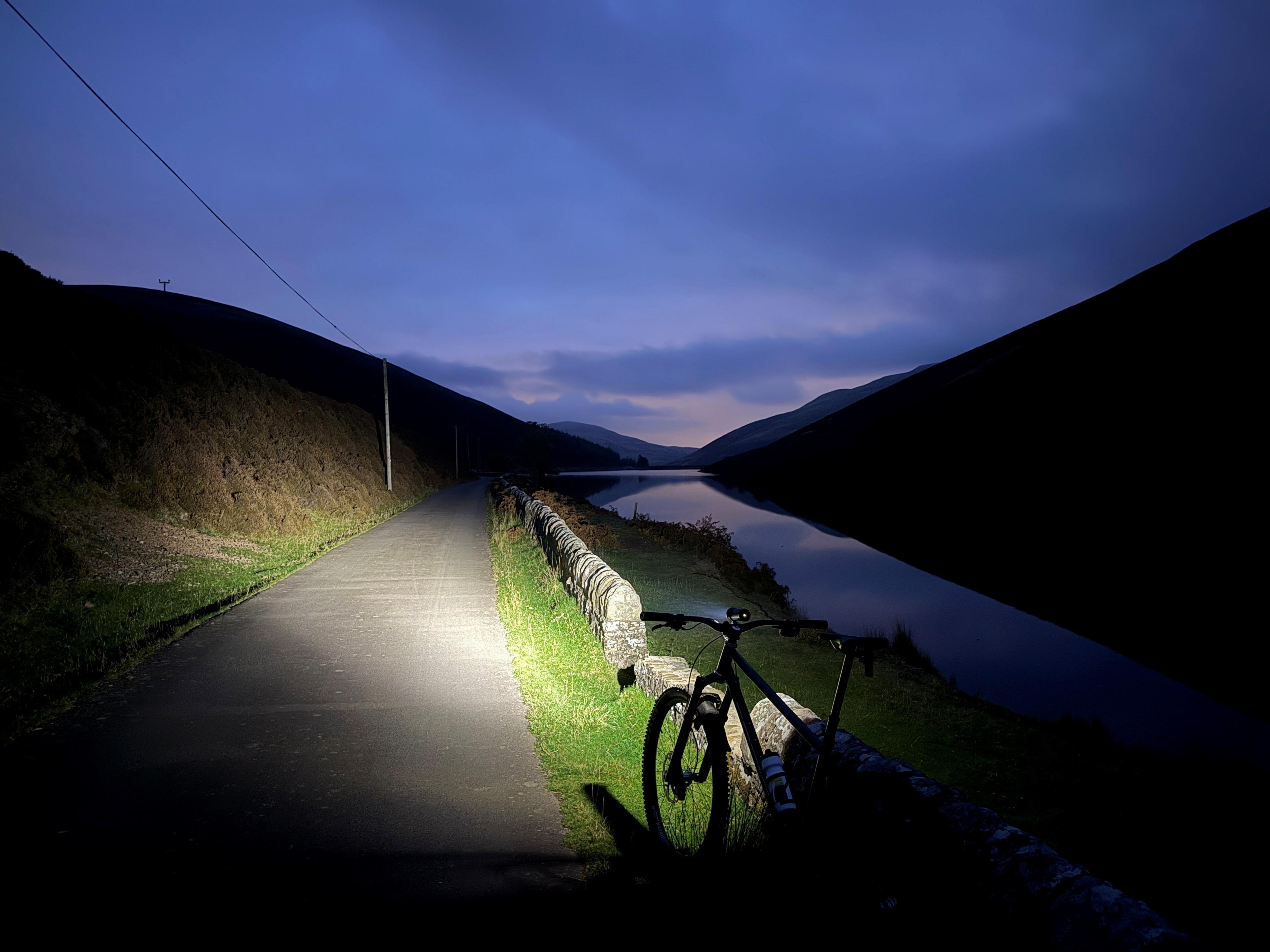 A bike with a light shining ahead o it. It is illuminating a path next to a reservoir on a dark night.