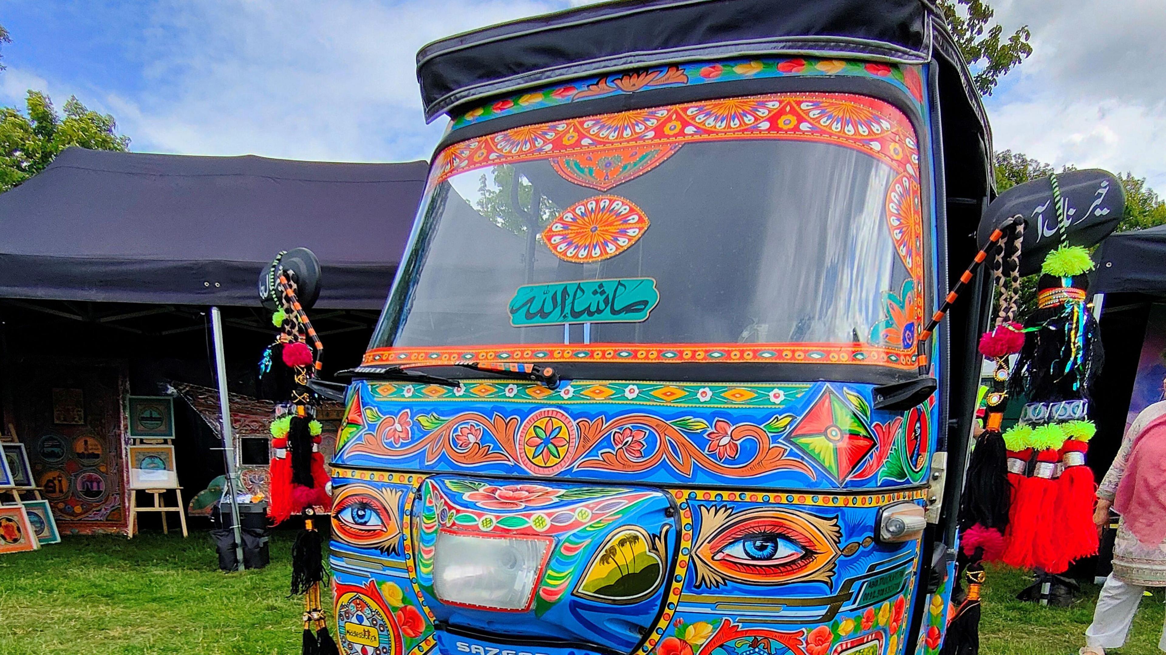 A brightly decorated auto rickshaw with colourful patterns and the words "The Leap" displayed on its windshield, There is a matching sign that sits on grass at an outdoor market or festival.