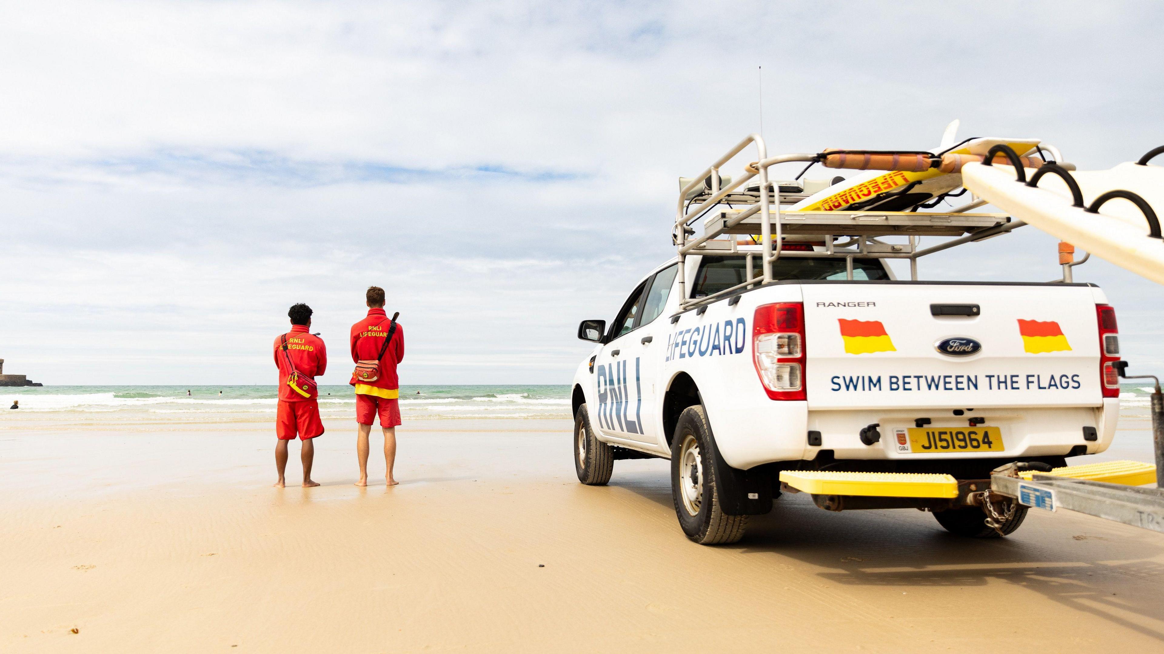 Two male lifeguards in red uniforms stand with their backs to the camera looking at the sea beyond. An RNLI branded Ford pick-up truck is parked next to them on the sand. It is loaded with paddleboards and reads 'RNLI LIFEGUARD' on the side and 'SWIM BETWEEN THE FLAGS' on the back.