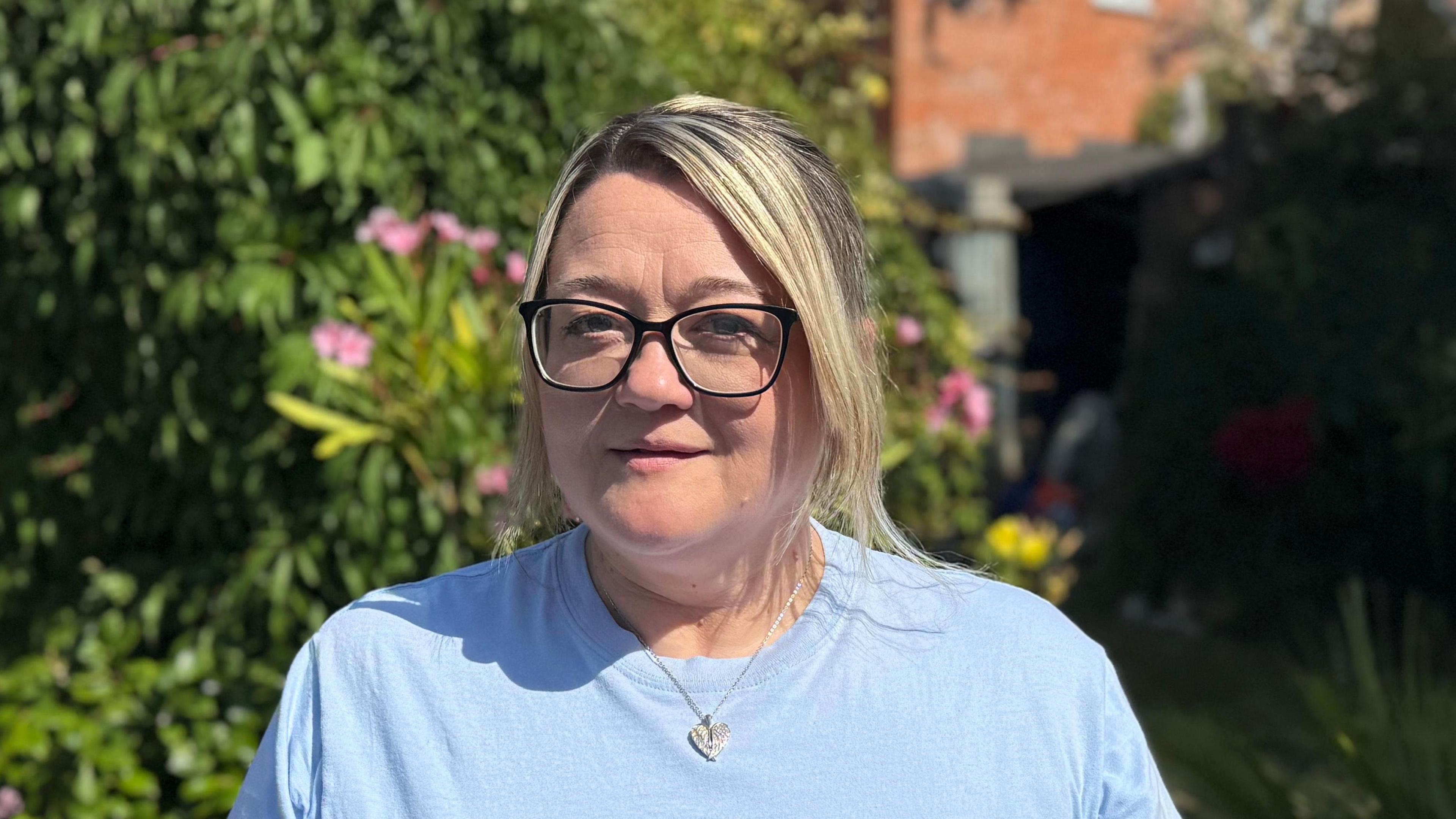Samantha Mitchell standing in her garden. She is wearing a pale blue T-shirt and glasses. She is looking into the camera.