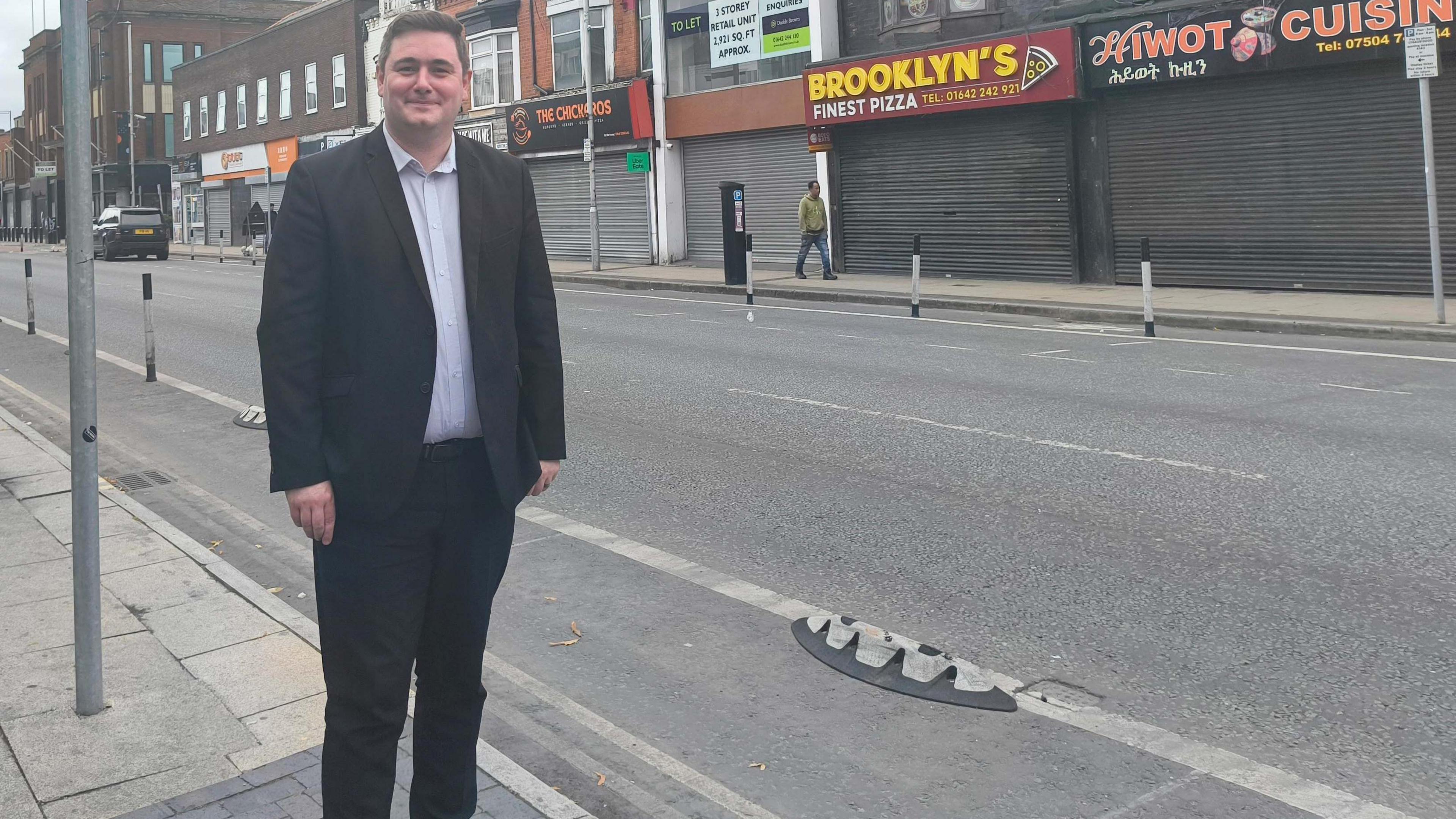 Mayor Chris Cooke, wearing a dark suit, is stood next to a cycle lane with a bin not far behind him. There are shops on the other side of the road.