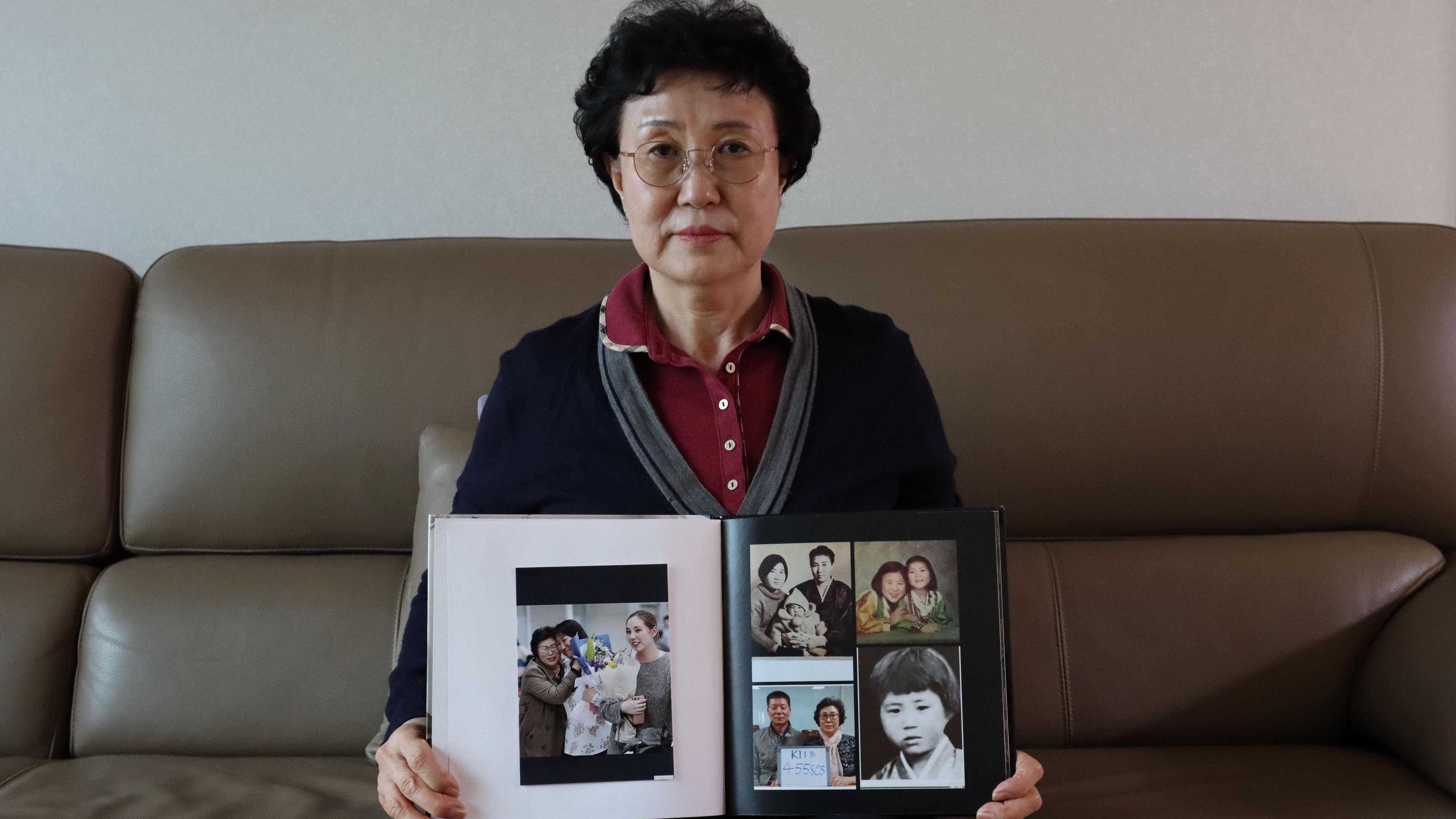 Han Tae-soon looks at the camera while sitting on a brown leather sofa, holding a photo book filled with pictures of her daughter Kyung-ha
