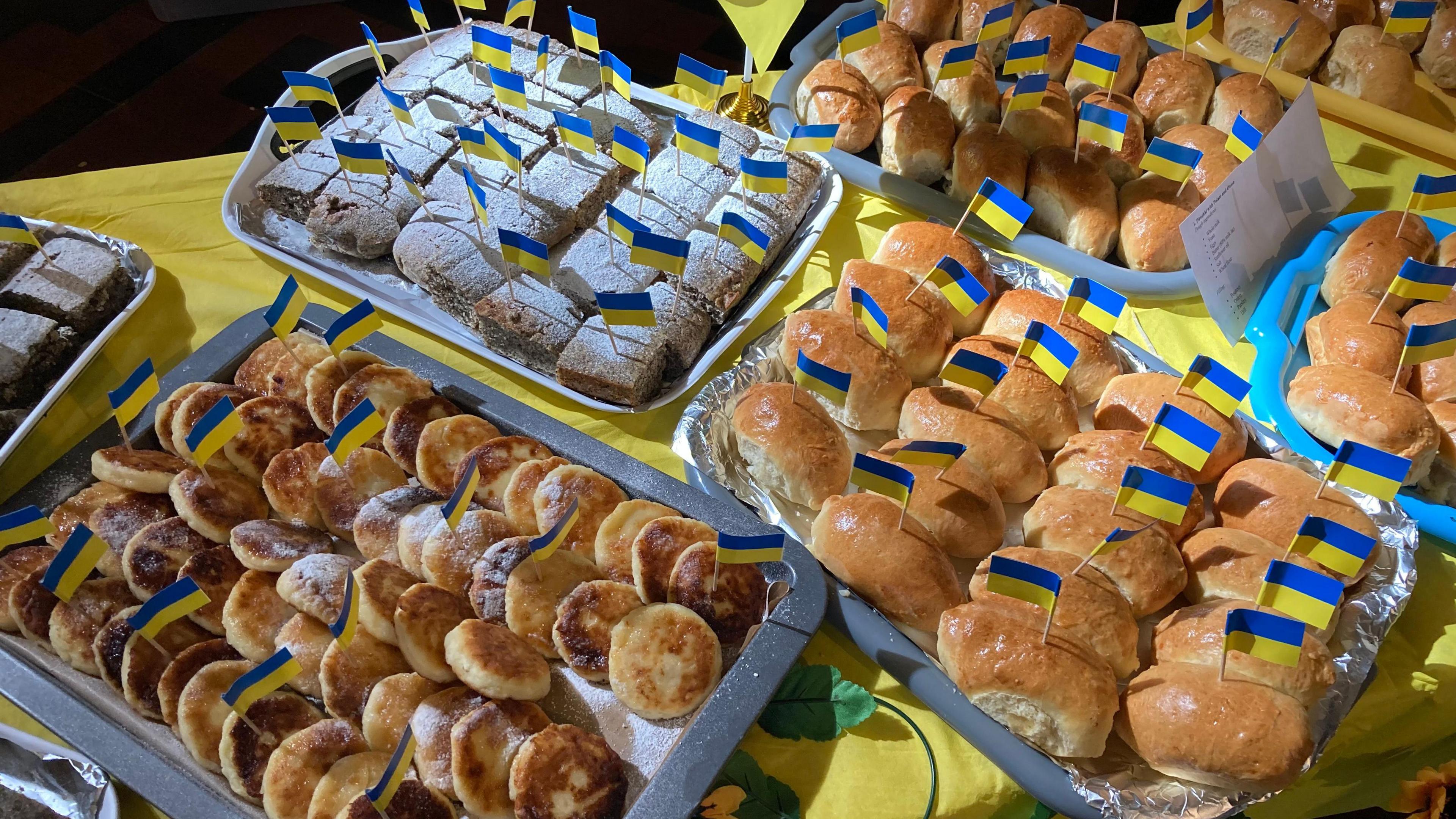 A selection of different Ukrainian cakes laid out on trays on top of a yellow tablecloth. Each cake has a stick with a Ukrainian flag.
