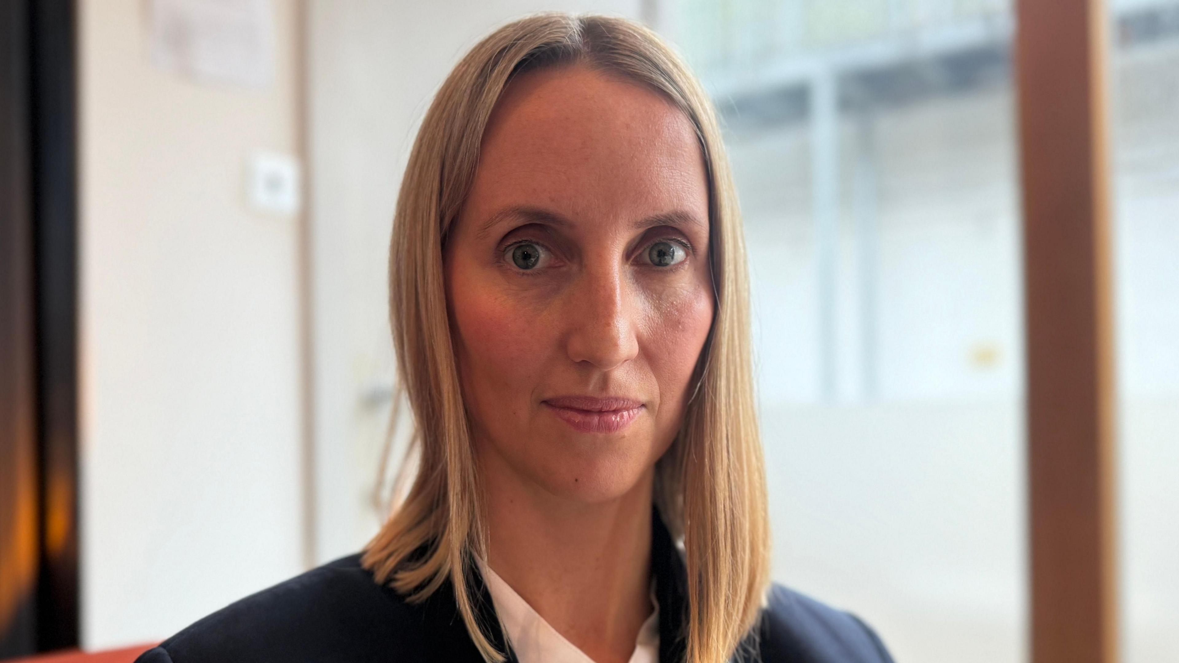 Sarah Woodcock has blue eyes and a blonde bob, she is wearing a blue jacket with a white top underneath. The background behind her is blurred.