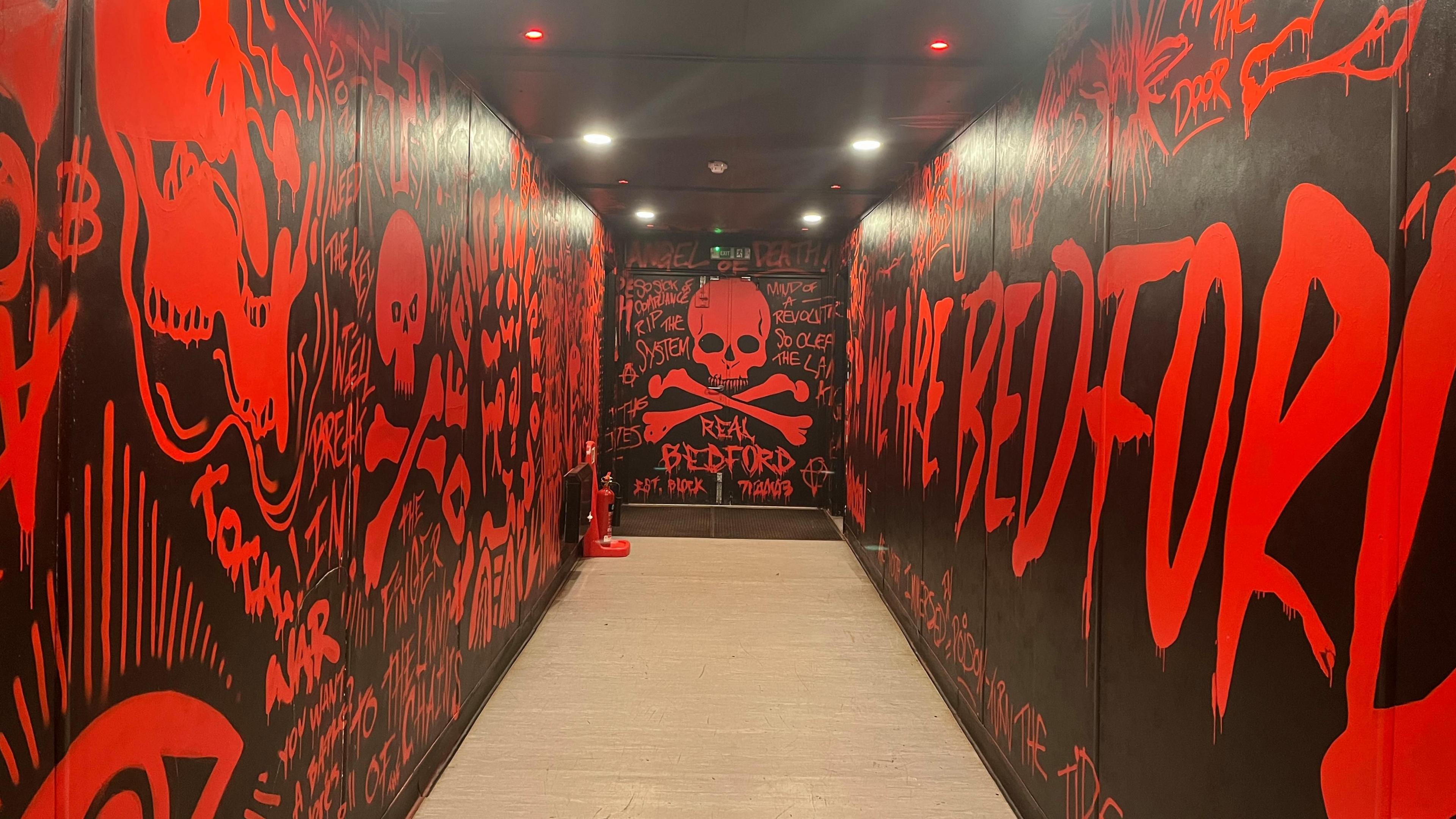 The players' tunnel at Real Bedford's football ground, with black walls and red lettering with a skull and crossbones at the end, and the words "Real Bedford".