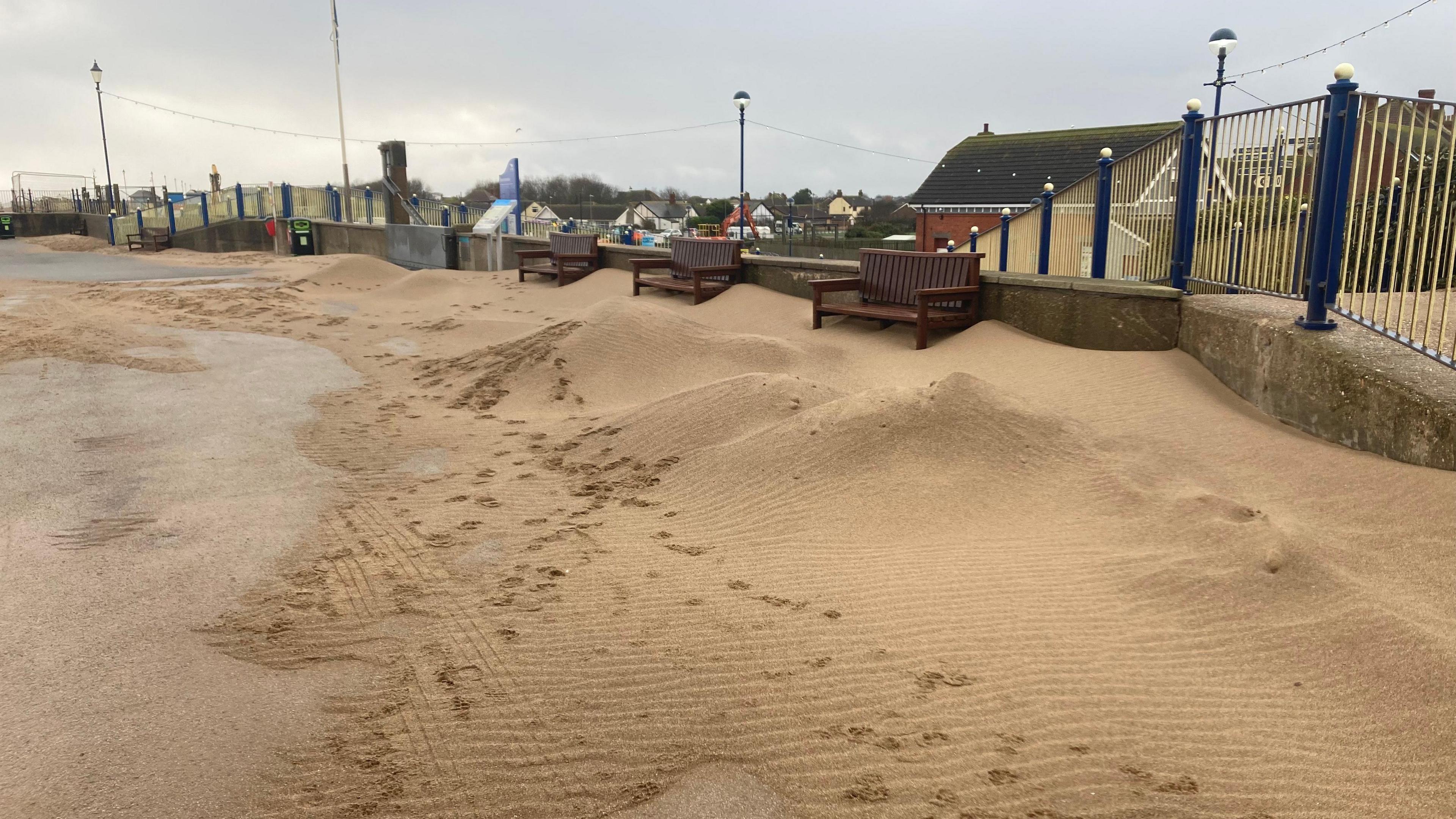The photo shows a beach walkway. To the right hand side are piles of sand, which are almost as high as the wall in the background which is running adjacent to the path. There are three benches just in front of the wall. They are blocked off by the piles of sand.