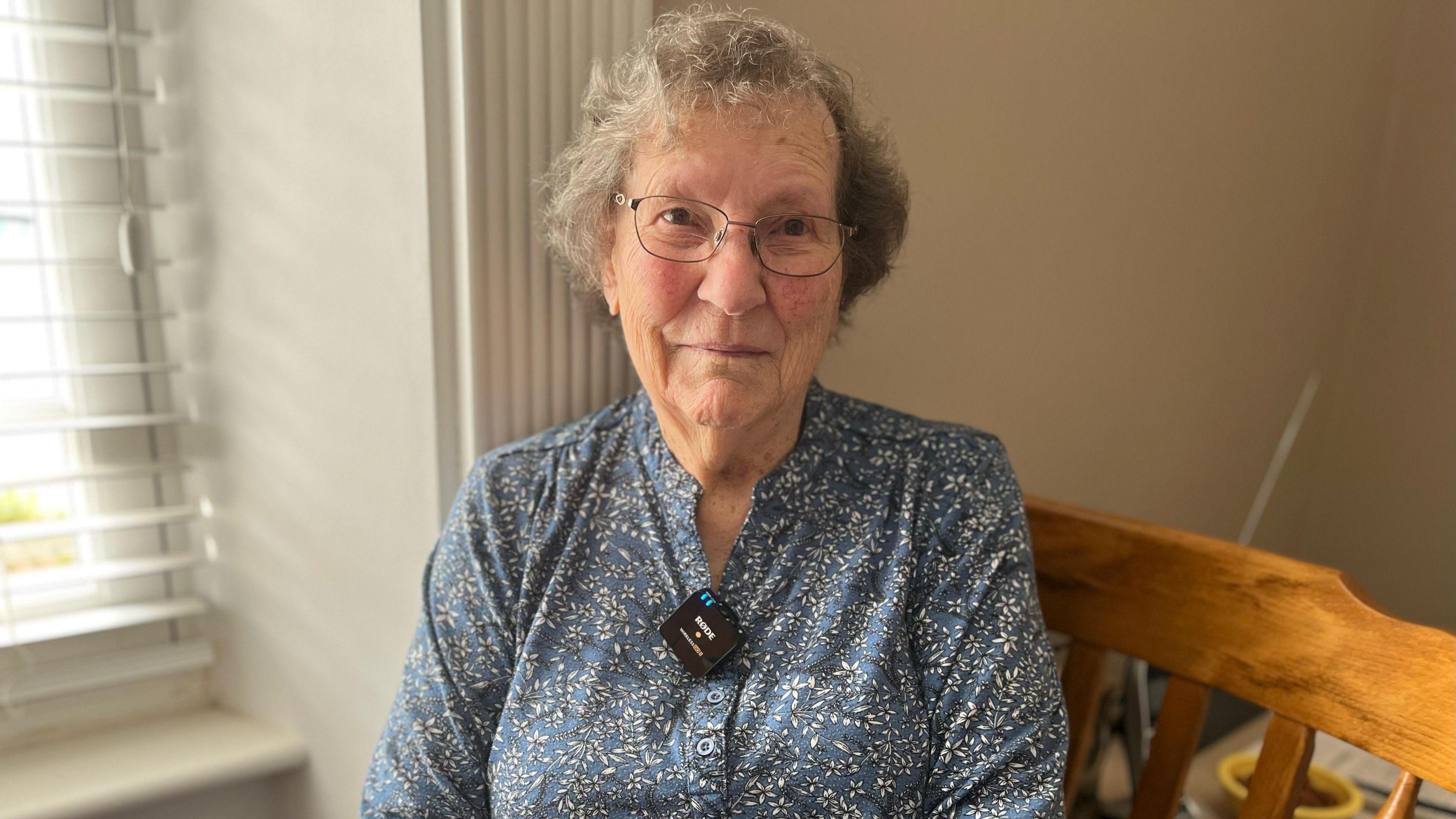 Lady with short grey hair, rosy cheeks and glasses, wearing a blue blouse sits on a  wooden chair next to a window.