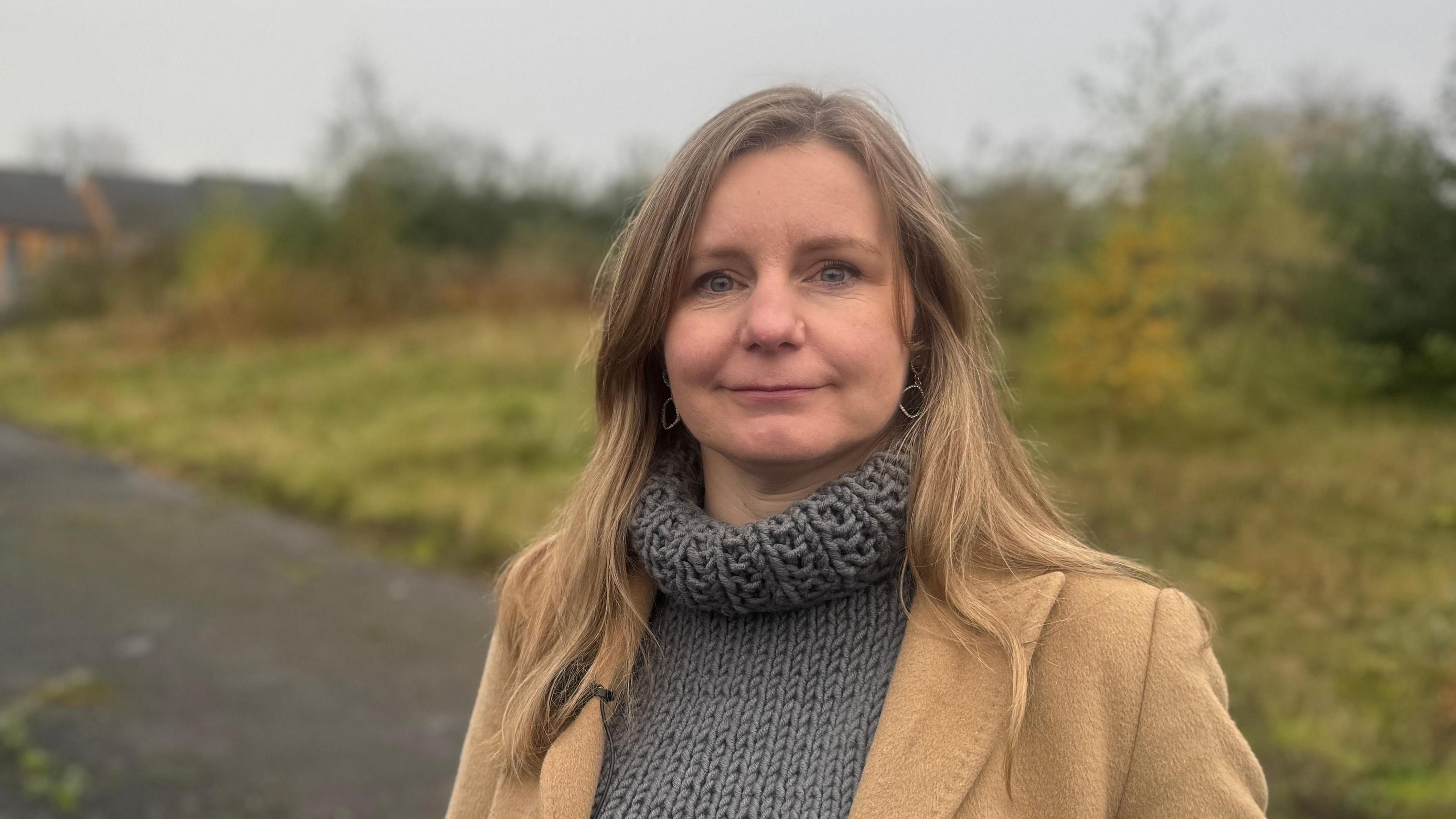 A woman wears a brown coat and grey turtle neck sweater. Behind her is a gravel path which sits in front of a green field.