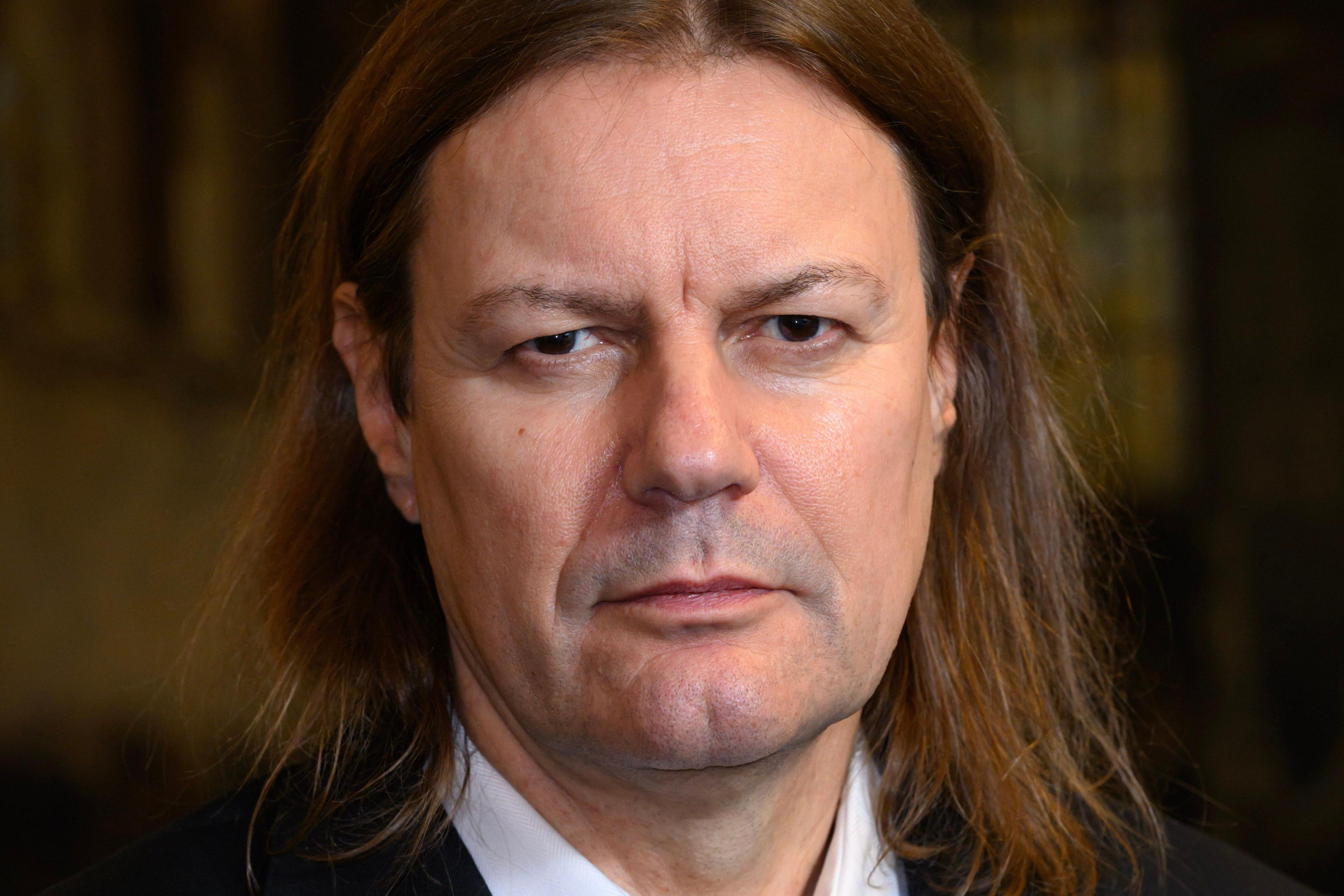 Mike Nicholas with long hair and wearing a dark suit looks into the camera 