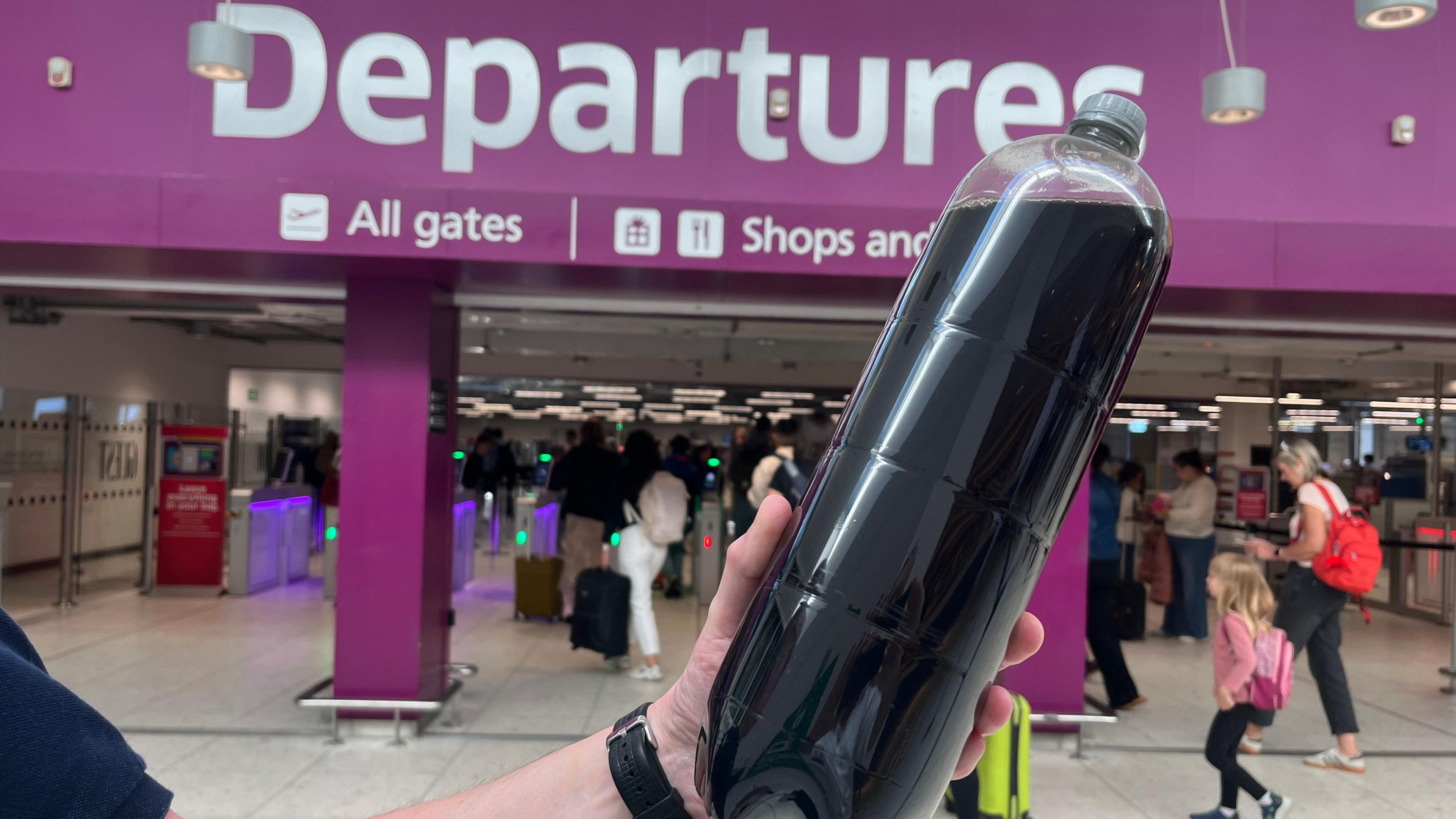 A 2 litre bottle in front of a purple sign that reads "Departures". People walk through the opening below the sign.