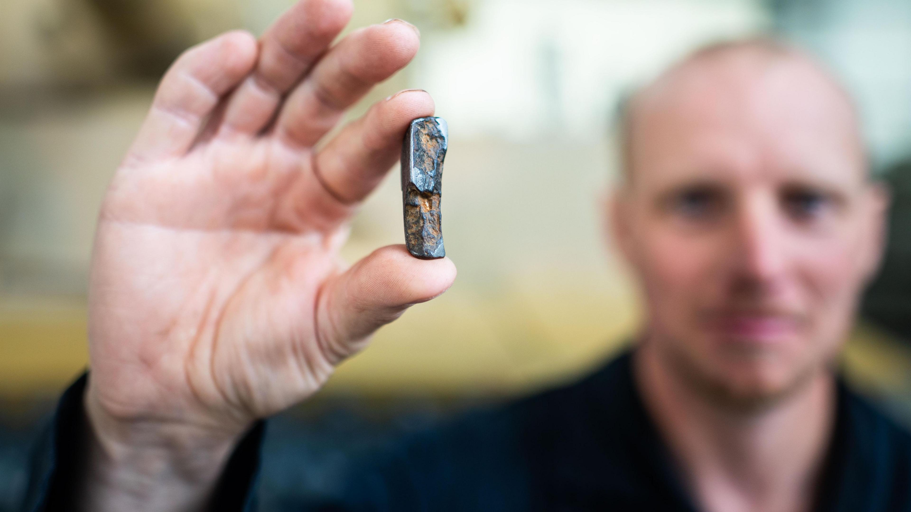 Blurred face of man with his hand held forwards - in focus - he is holding a piece of gnarled shrapnel between his thumb and finger.