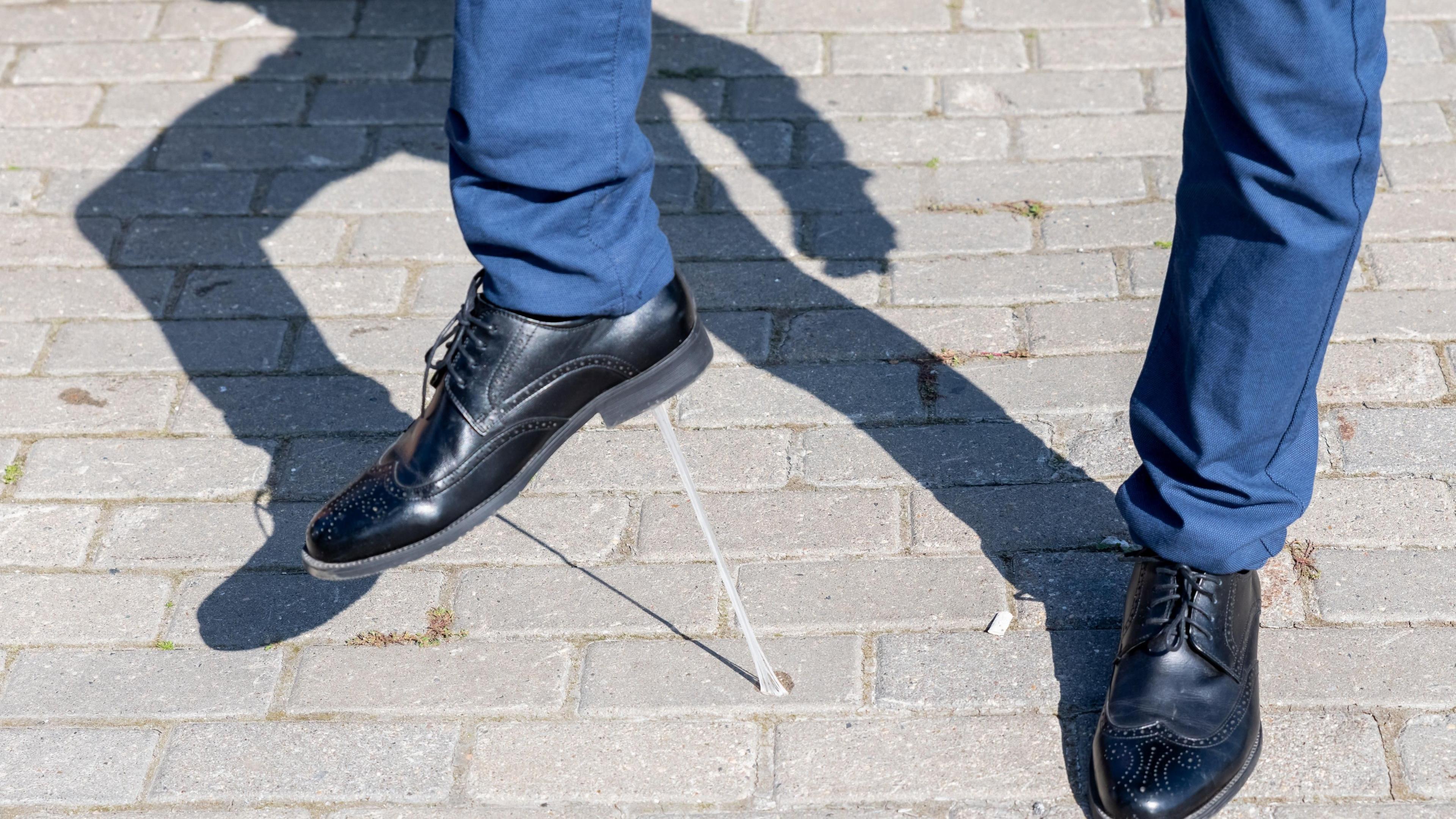 A stock photo showing the bottom half of a pair of legs in blue trousers and smart black shoes. The person is lifting up their right foot - they have stepped in gum, which is now stuck to the bottom of their shoe.