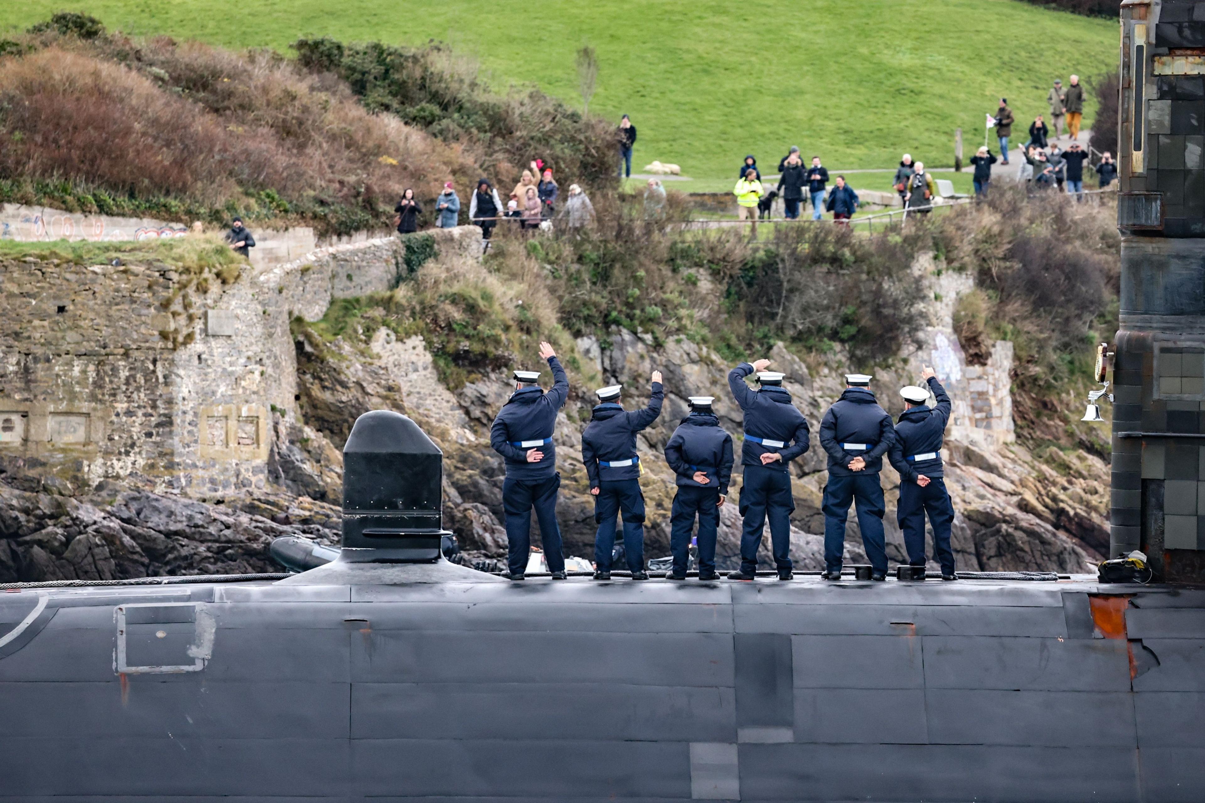 The submarine arriving at port, with five personell on deck in white hats seen from the back waving to people on a rocky and grassy shoreline.