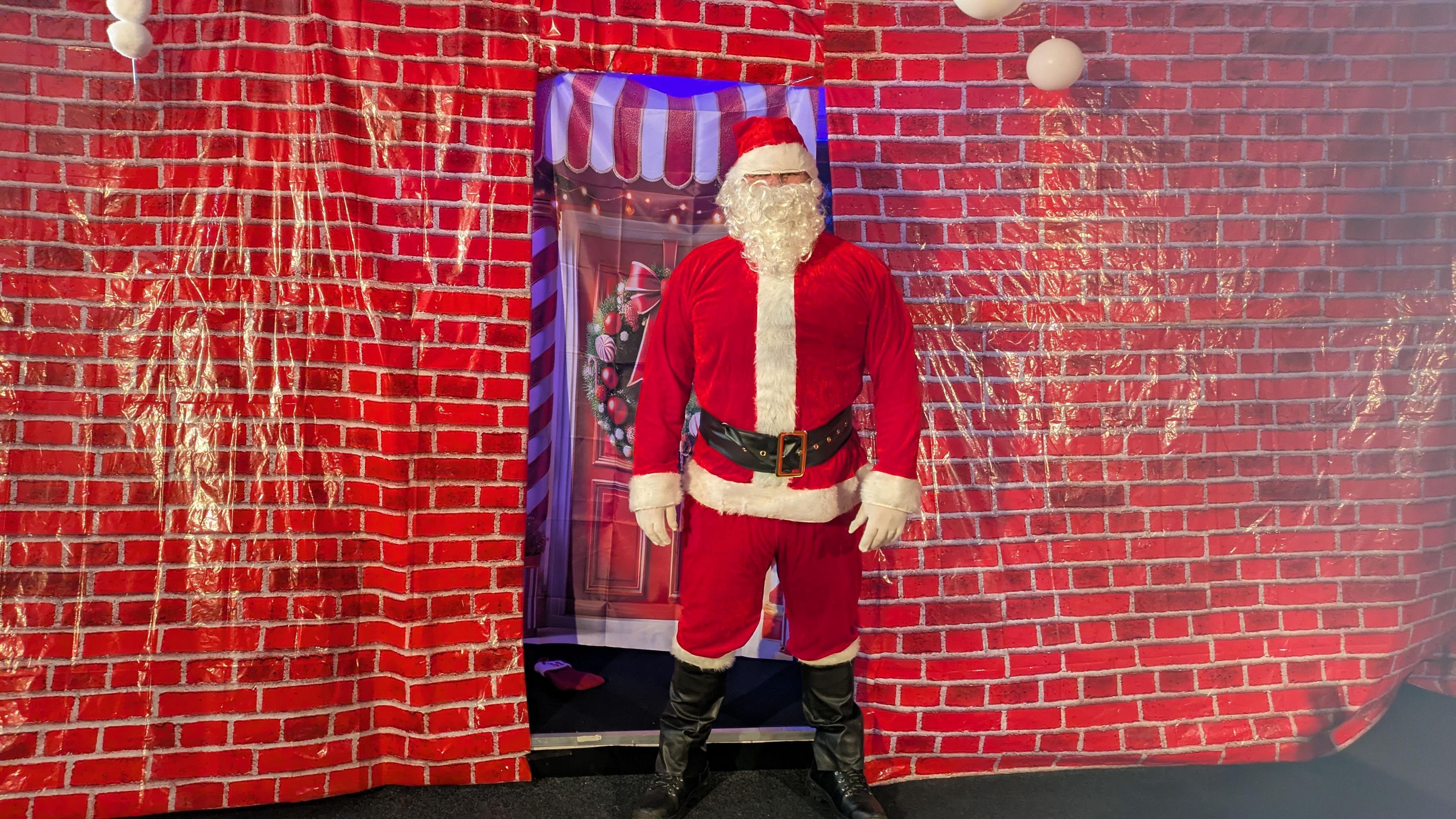 A man dressed in a Santa Claus suit and black boots, standing in front of sheeting made to look like a red brick wall with a festive door.