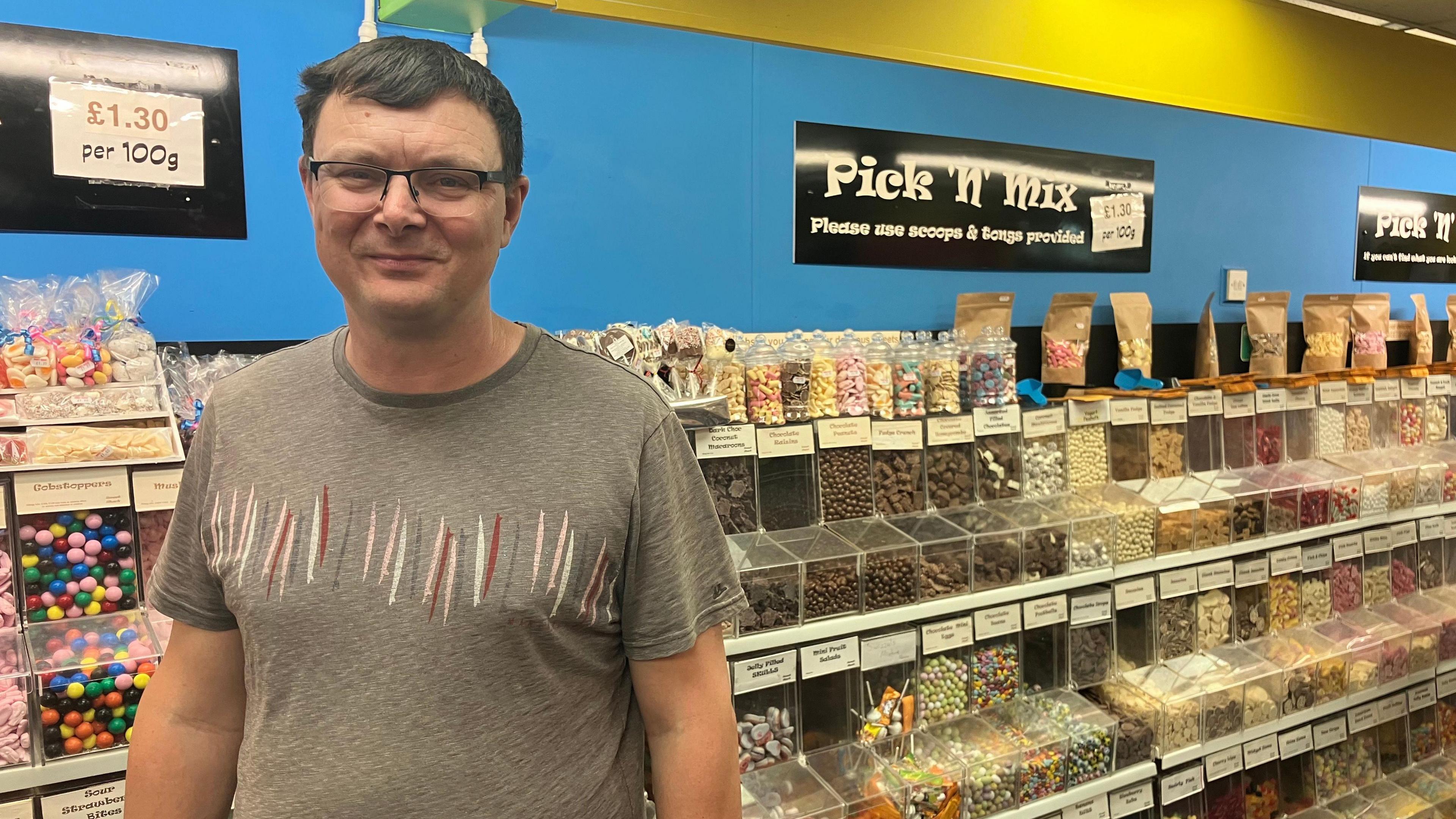 John Mills standing in front of a pick and mix stand in a shop, He is wearing a grey T-shirt and glasses