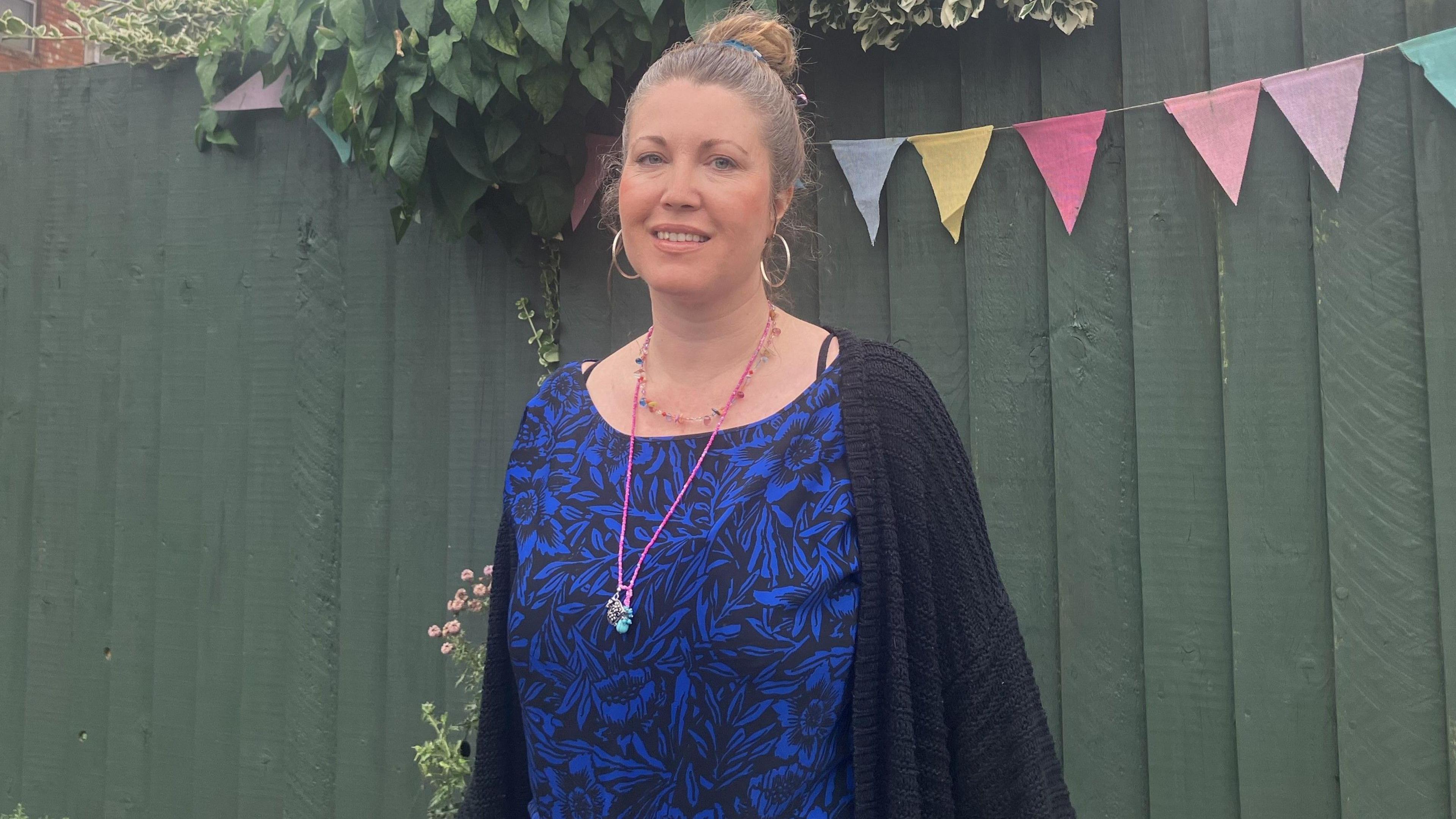 A woman in a blue dress stands in front of a sage green fence