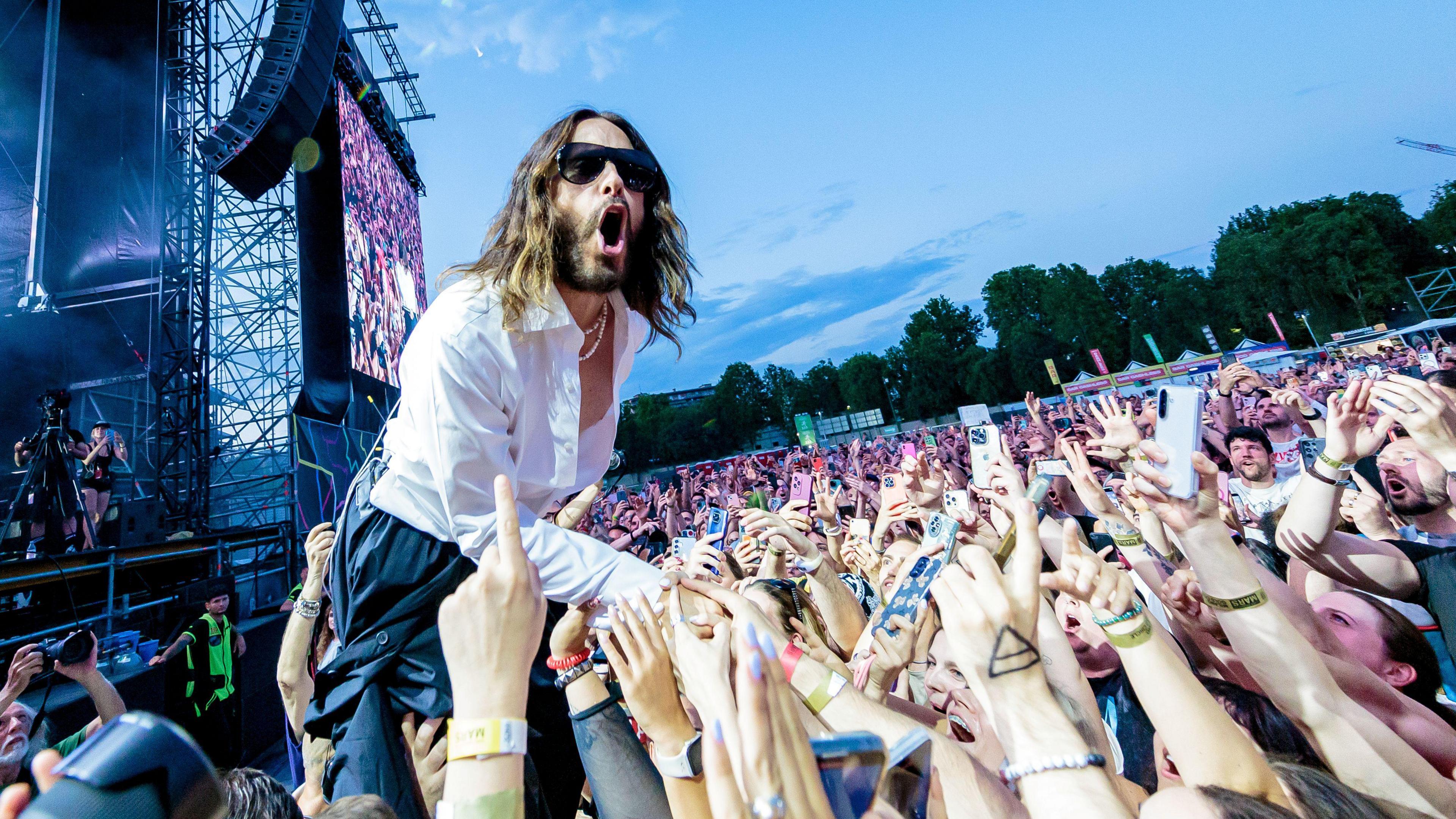 Jared Leto performing at a concert. He wears a white shirt and is leaning into a sea of fans who have their arms raised up.