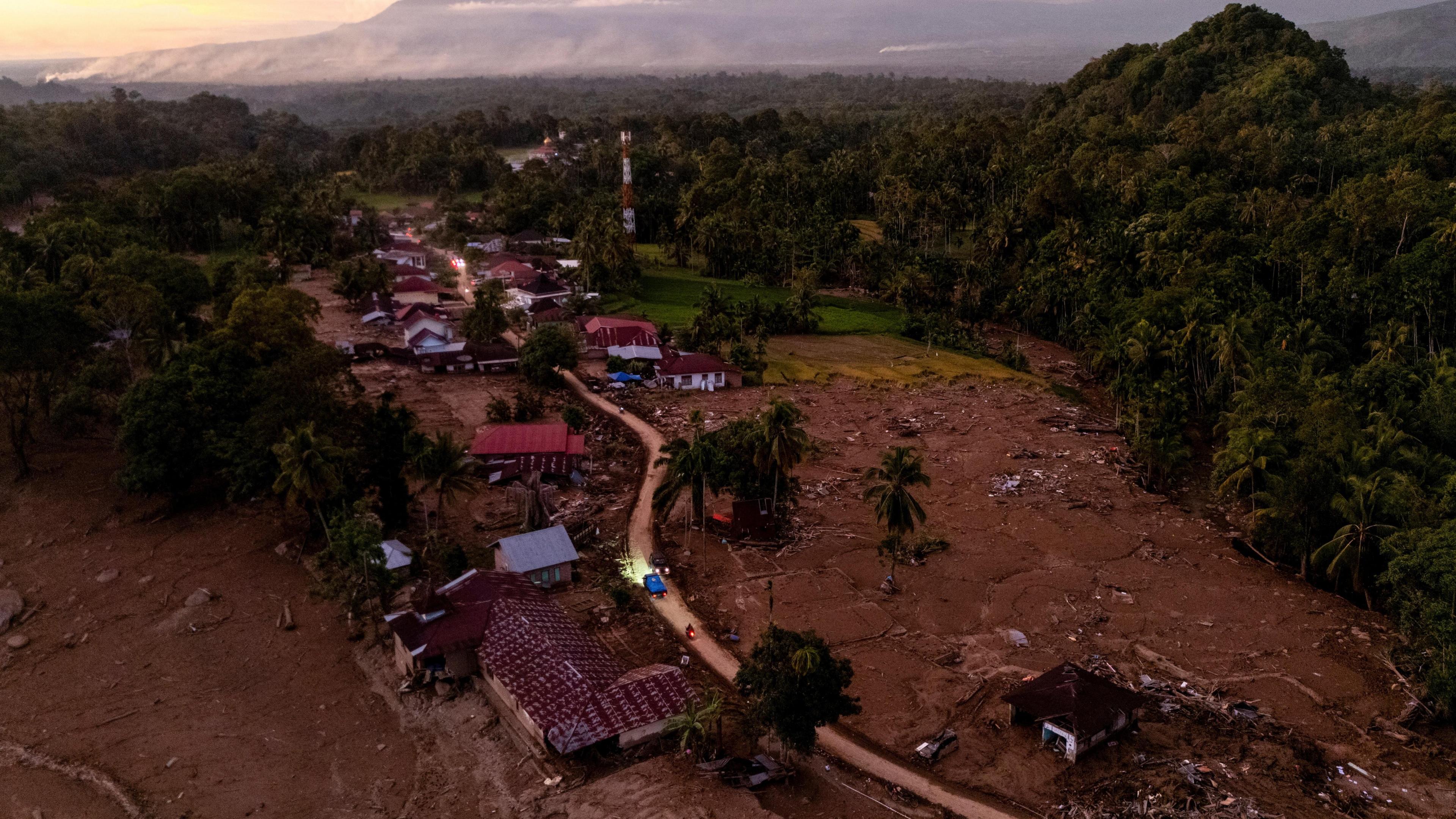 A drone view of vehicles driving through an area hit by deadly flash floods following heavy rains in Palembayan, Agam regency, West Sumatra province. The road cuts through fileds which appear to have been turned to mud, the houses are damaged. The area is surrounded by jungle, with a mountain in the background