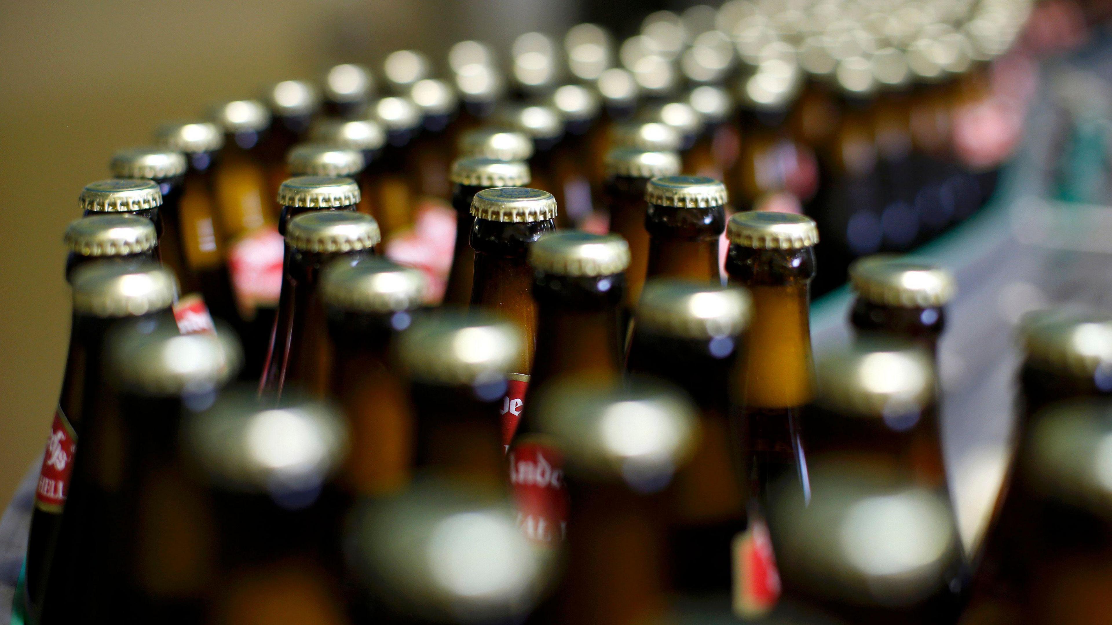 Beers bottles lined up on a conveyor belt at a factory. They are brown with gold caps.