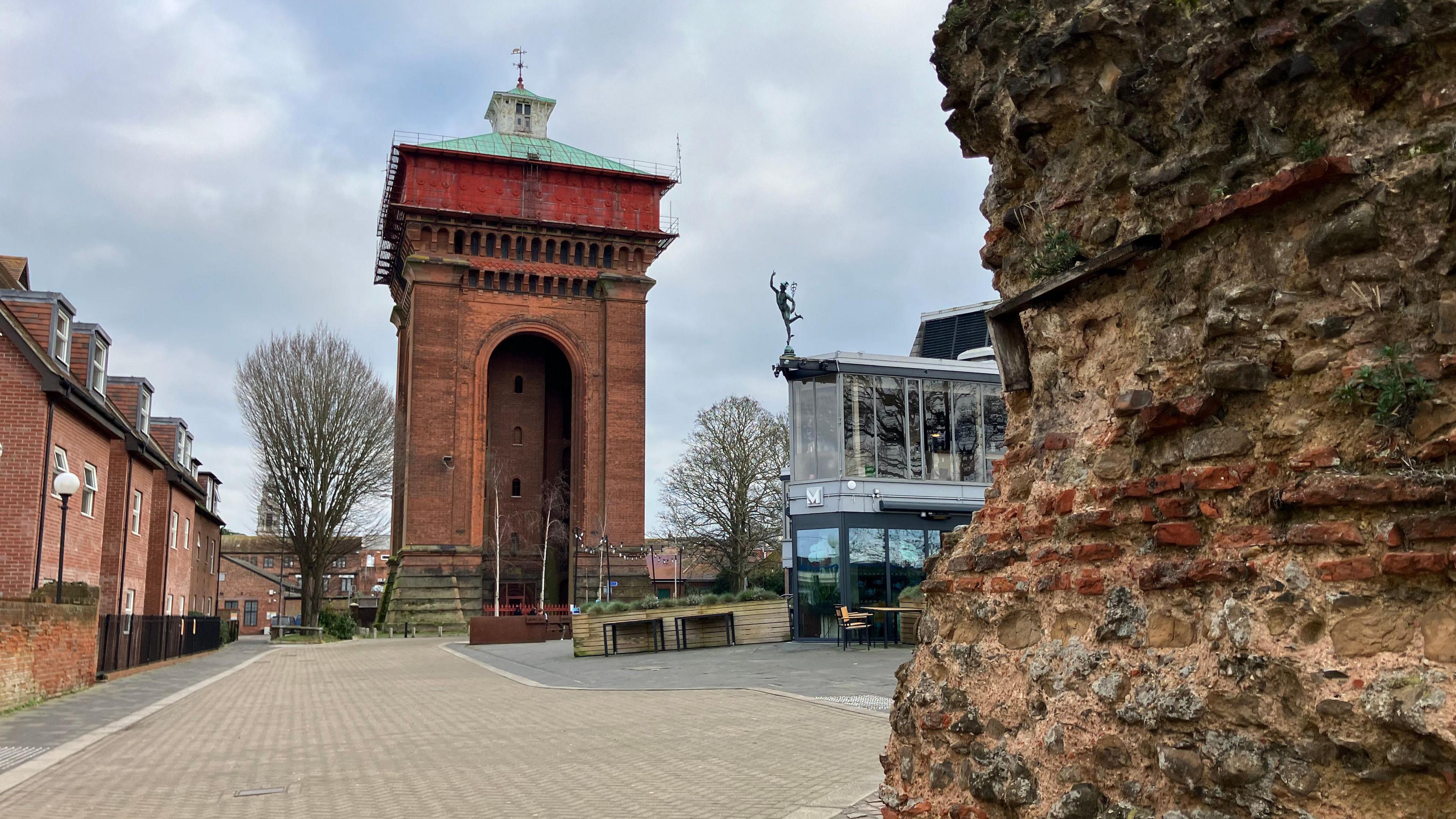 A large, red brick water tower stands in a public square. It has a green roof and a rust-coloured section at the top. Also pictured is the side of a Roman wall, and a modern glass building which houses the Mercury Theatre.