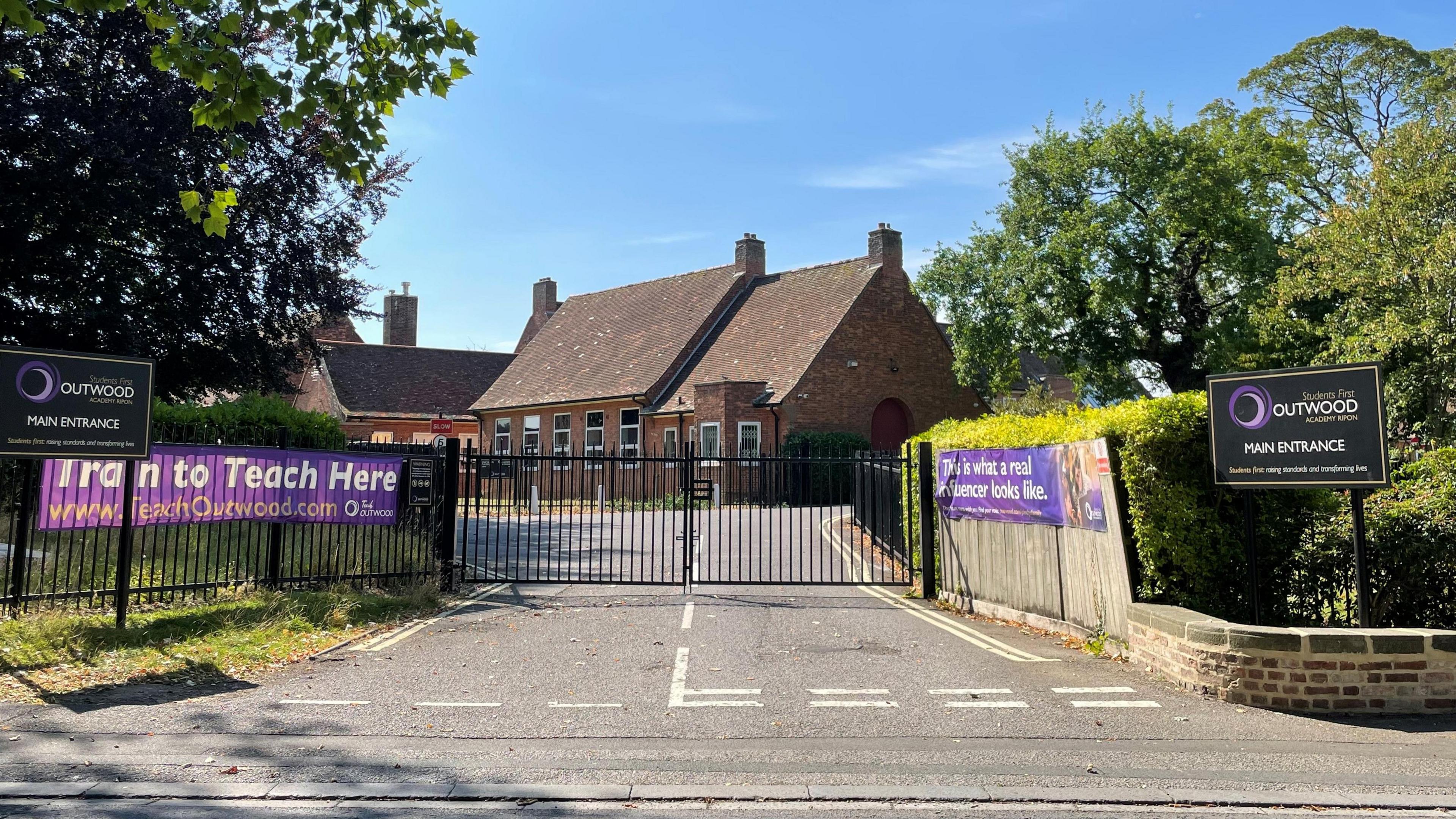 A school behind closed black gates. There are purple promotional signs which invited people to "Train to Teach Here" and "This is what a real influencer looks like."