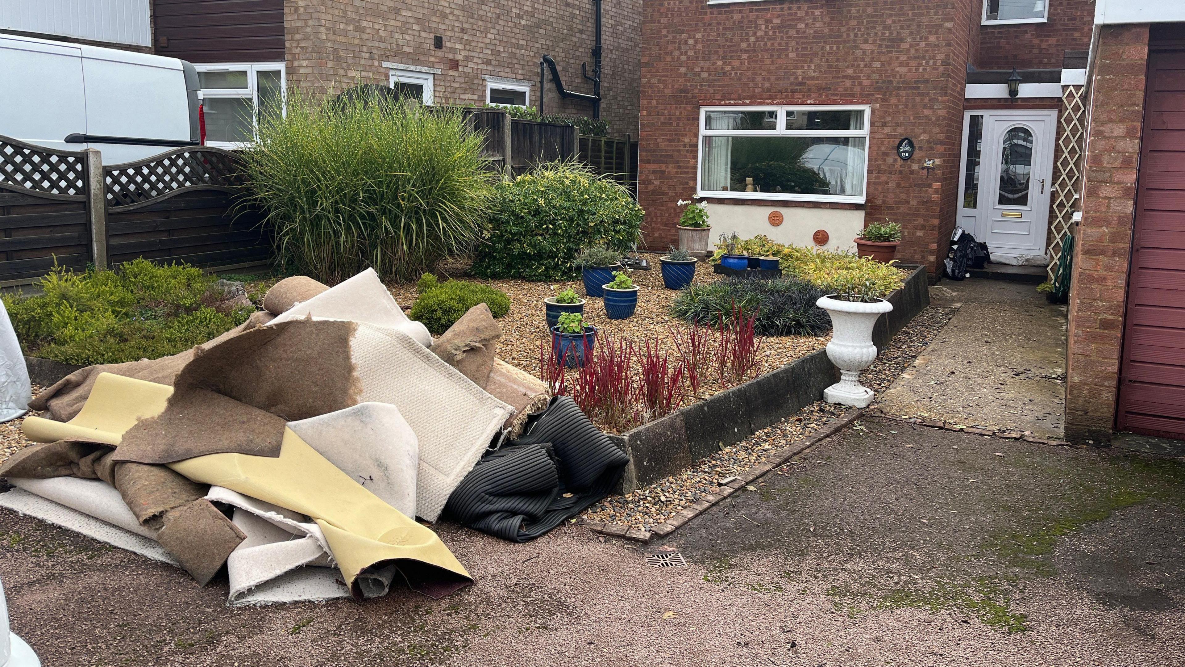 A pile of carpets and underlay lying on the pavement at the end of a house's front garden. The carpets are partially rolled and folded up, they are light brown, yellow, black and cream in colour. The front garden is mostly gravel, with blue plant pots, low level flower beds and a couple of larger shrubs. A tarmac and concrete path on the right of the image leads up to a white front door. The house is brick built and one large ground floor window with a white frame can be seen in the background. 